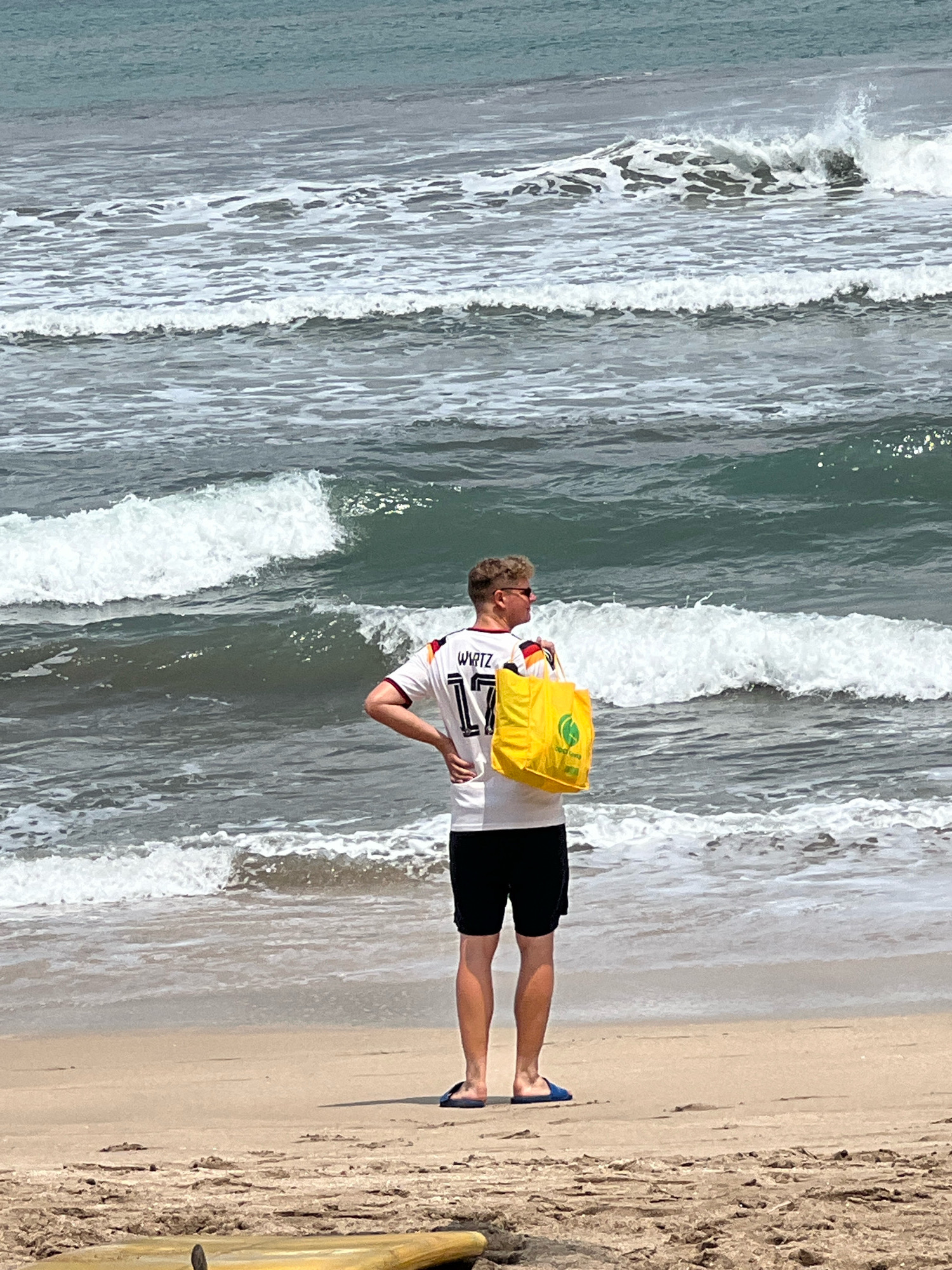 A person stands on a sandy beach, facing the ocean. The person is wearing a white jersey with the number 17 and the name "WIRTZ" on the back, and black shorts. They have a yellow bag slung over their right shoulder. The ocean has several waves breaking, with foam visible. The beach sand has some debris scattered on it, and in the foreground, part of a yellow surfboard is visible. The sky is overcast, and the water appears to be a murky blue-green. The location is the Kuta beach in Kuta, Indonesia.