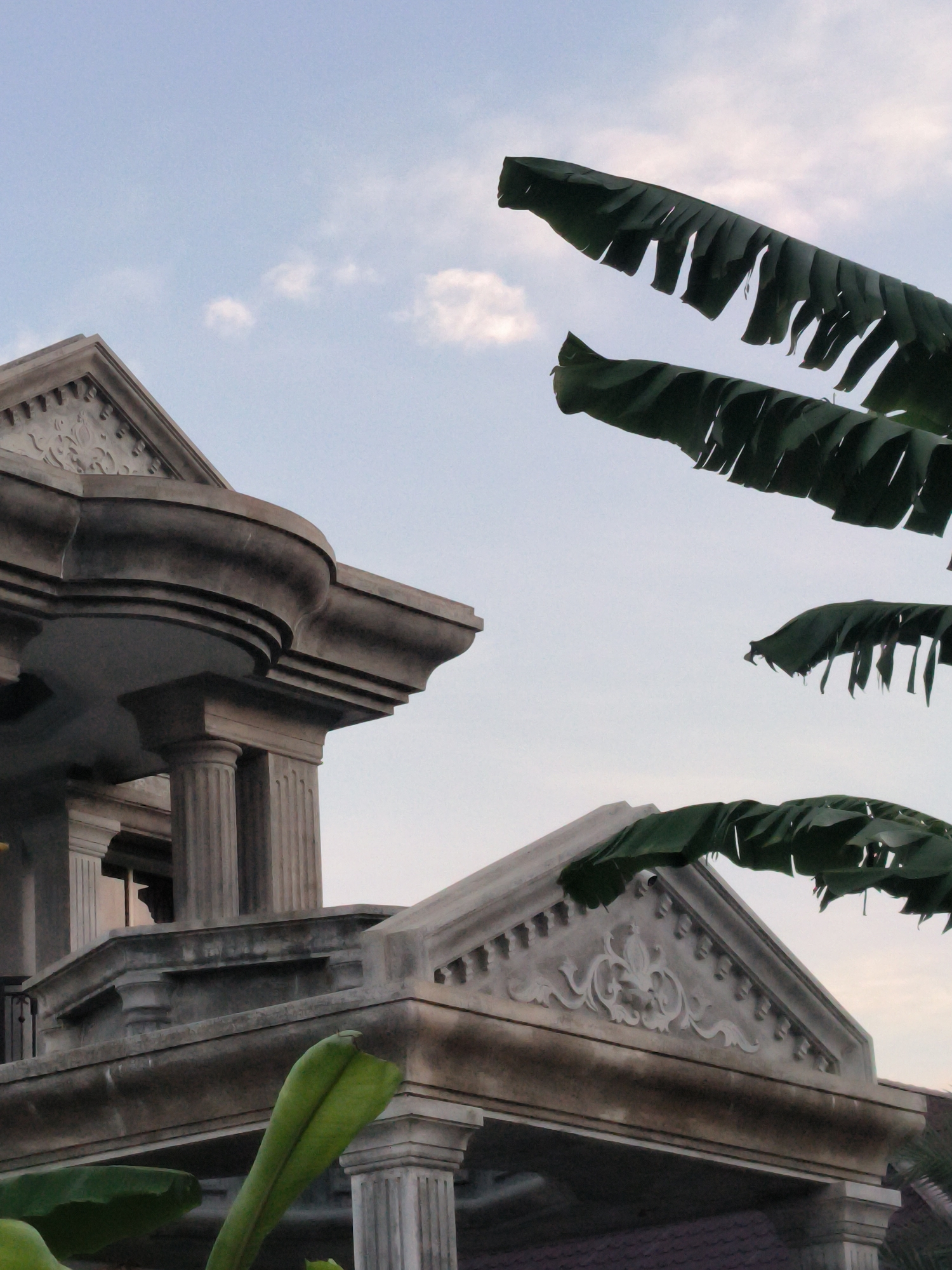 The image depicts a partially obscured view of an ornately decorated building exterior, with elements suggesting a neoclassical or Beaux-Arts architectural style. Details include pillars with fluted shafts, elaborate cornice work, and decorative relief carvings on the pediment. The building's color is a pale stone or concrete. The sky is light blue with some clouds, suggesting either morning or late afternoon. In the foreground, there are large, green banana leaves, partially framing the building, and lending a tropical feel to the scene. The location is Lhokseumawe, Indonesia.