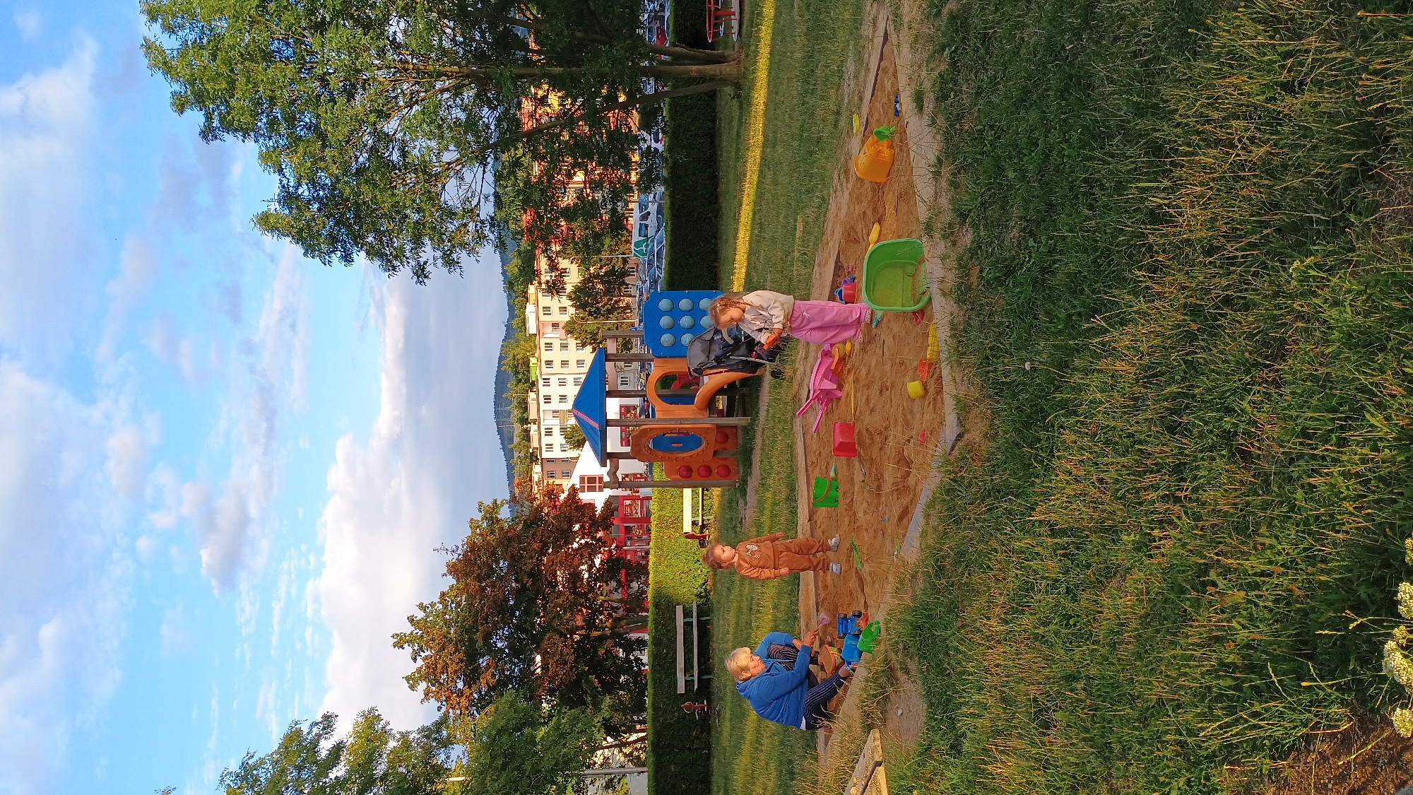 The image shows a playground in Sokolov, Czechia, under a partly cloudy sky. In the foreground, a sandpit is filled with children's toys, including buckets, shovels, and a toy truck. Two young children, a boy in a brown outfit and a girl in pink pants and a cream-colored top, are playing in the sand. An adult, likely a grandparent, is also in the sandpit, sitting and playing with the children. In the background, a playground structure with slides and climbing elements is visible, along with apartment buildings and trees. The setting appears to be a residential area, possibly a park or communal space for residents. The sun is low in the sky, casting long shadows and illuminating the scene with a warm, golden light.