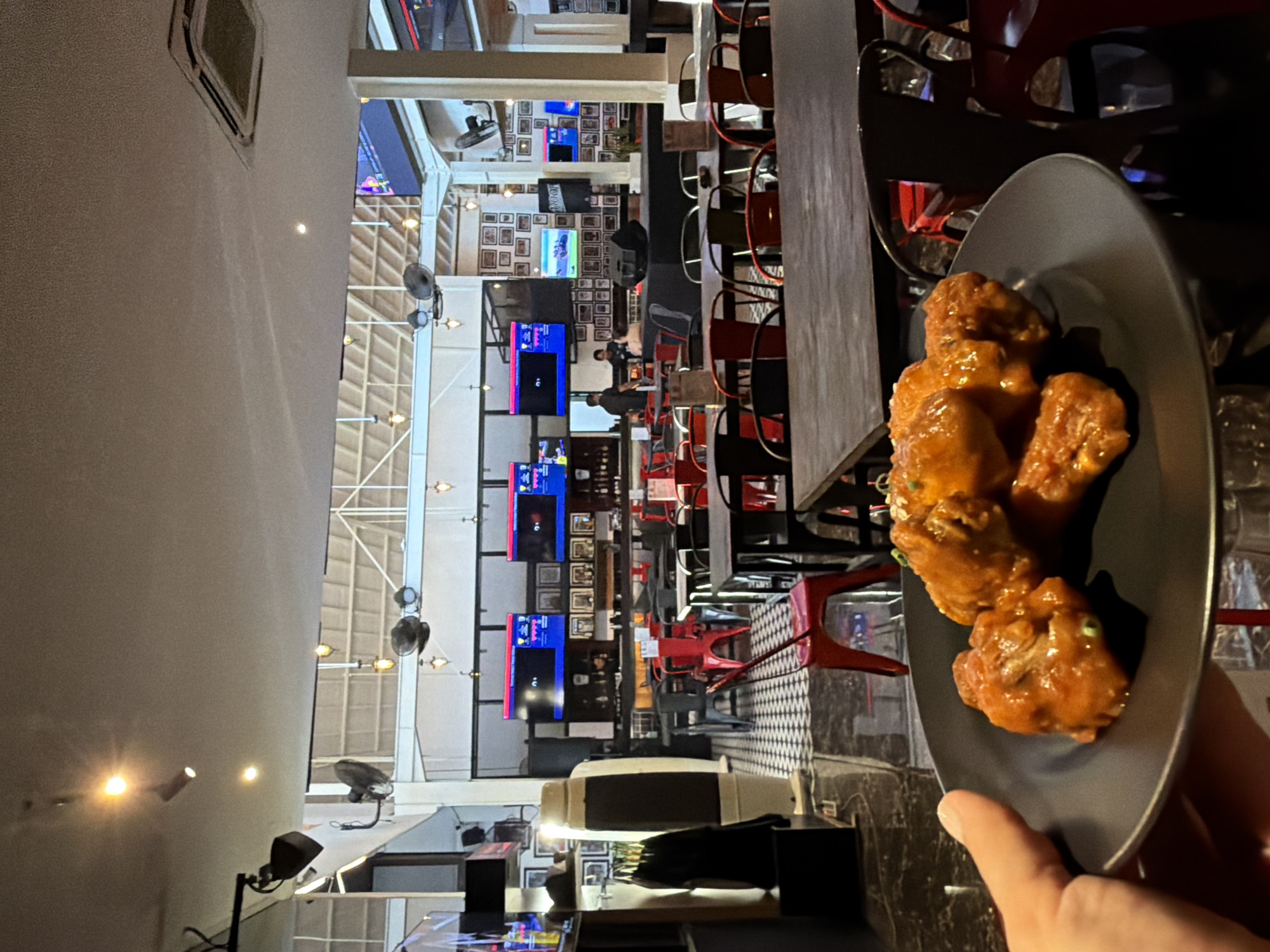 This media file presents an indoor scene, likely a sports bar or casual restaurant in Kuta, Indonesia, characterized by a lively and open atmosphere.
**Subjects:** In the immediate foreground, a person's hand is visible, holding a plate of food. In the mid-ground and background, several other individuals, likely patrons or staff, are scattered throughout the establishment, some seated at tables and others standing.
**Objects:** The most prominent object is a dark grey or black plate held in the foreground, laden with several pieces of chicken wings. The wings are coated in a thick, glossy, reddish-orange sauce and appear to be garnished with fine green specks, possibly chopped scallions or herbs. The background is replete with numerous flat-screen televisions mounted on various walls and pillars, displaying diverse visual content, including what appears to be sports or entertainment. The walls between the screens are decorated with a dense grid of framed pictures or artworks. The interior is furnished with many tables and chairs; some chairs are distinctively red, while others are dark-colored bar stools. Parts of the flooring feature a black and white checkerboard pattern. Ceiling fans, spotlights, and other light fixtures illuminate the expansive space, which also showcases exposed structural beams and high ceilings. A dispenser-like object is visible on a counter in the mid-ground.
**Scene/Setting:** The setting is a spacious, modern, and industrial-chic establishment, typical of a sports bar or family-friendly restaurant designed for dining and entertainment. Its open layout with high ceilings creates a dynamic and communal environment.
**Activity or event taking place:** The scene depicts patrons dining and socializing. The presence of multiple large screens suggests watching sports or other media is a central activity. The overall ambiance indicates a casual gathering or meal.
**Notable details:** The lighting is entirely artificial, consisting of spotlights and ambient fixtures, strongly suggesting that the time of day is evening or night. The multitude of screens points to a venue that prioritizes broadcasting events or entertainment. The general mood appears informal and relaxed.
**Visible text:** On one of the television screens located towards the middle of the frame, the text "100%" is partially legible. Other screens display various graphics, but no other specific text is clearly discernible.