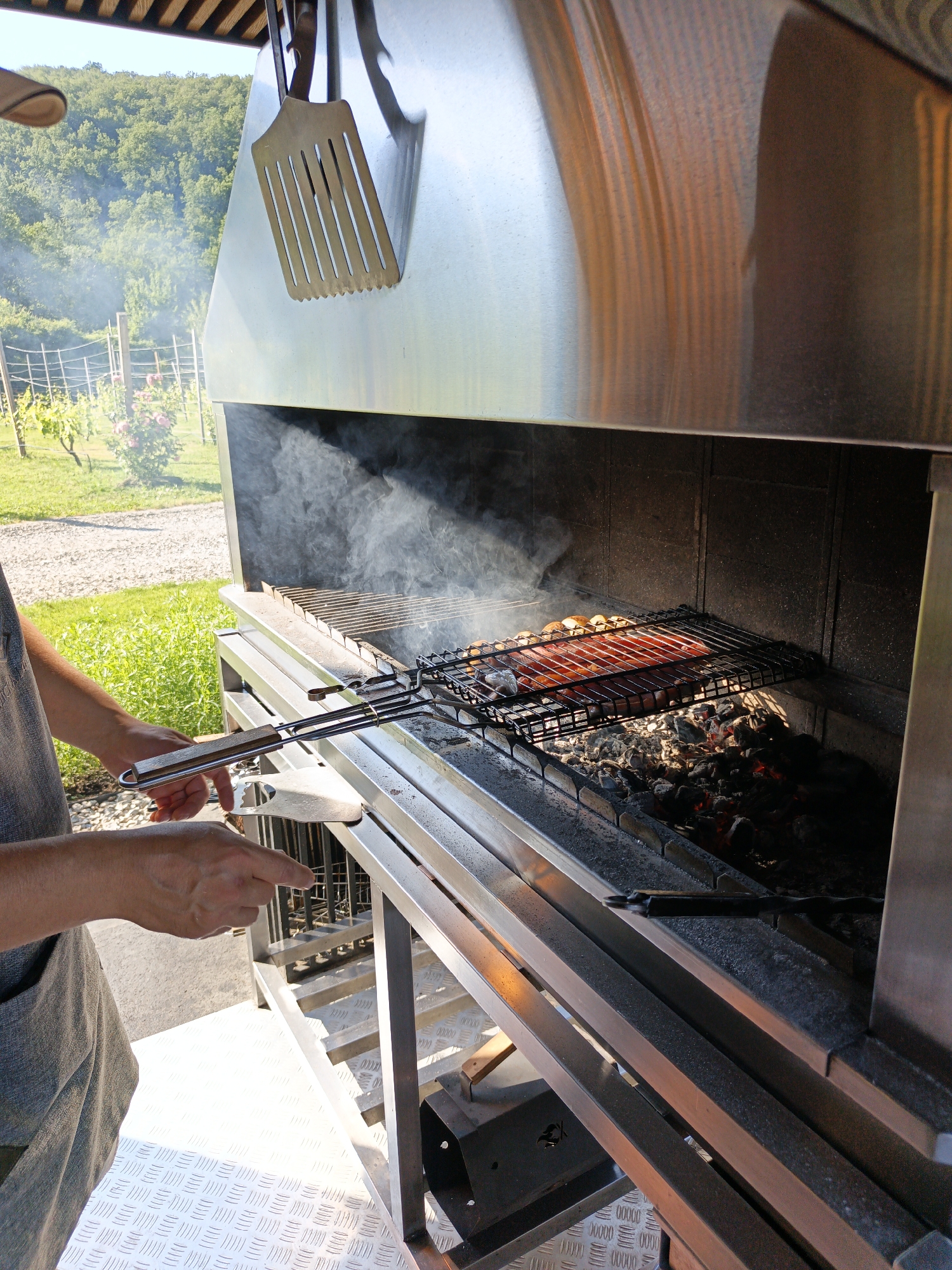 A person wearing an apron is grilling sausages and other food on a metal grate over hot coals. The grill is outdoors, under a covered structure. In the background, there are trees, a vineyard, and a blue sky. The scene appears to be in the city of Pshada, Russia.