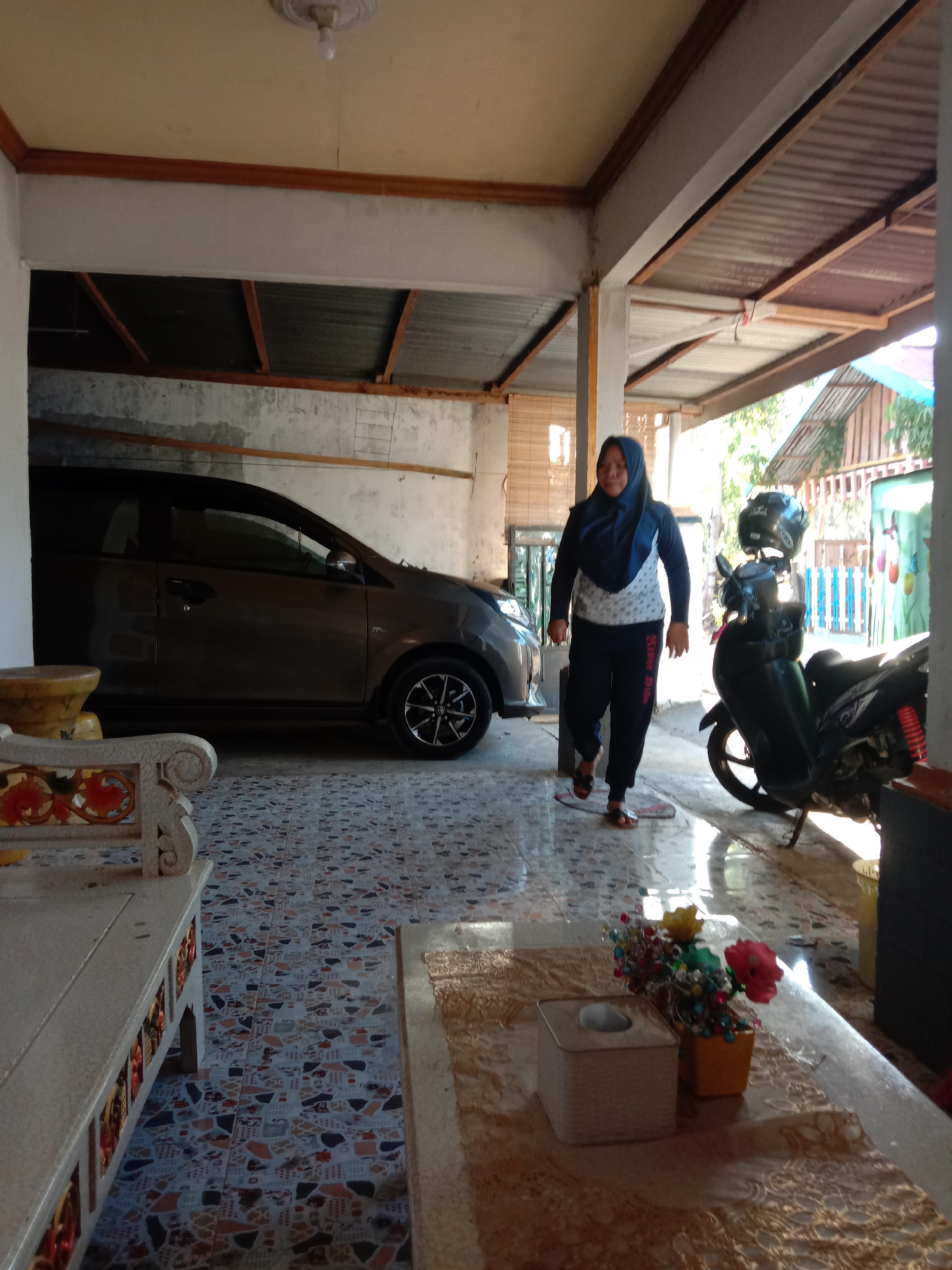 The image captures a scene in a covered outdoor area, possibly a carport or patio, in Kwandang, Indonesia. A woman wearing a dark blue hijab and a patterned long-sleeved shirt and dark pants is walking across a tiled floor. To her left, a dark-colored car is parked. To her right, a dark-colored motorcycle is parked, with a helmet resting on its handlebars. In the foreground, a long, light-colored bench with decorative carvings is visible, and a table with a tissue box and a small vase of artificial flowers sits on a patterned runner. The environment appears to be residential, with corrugated metal roofing and wooden beams visible overhead. The lighting suggests it is daytime, with sunlight casting shadows on the floor.
