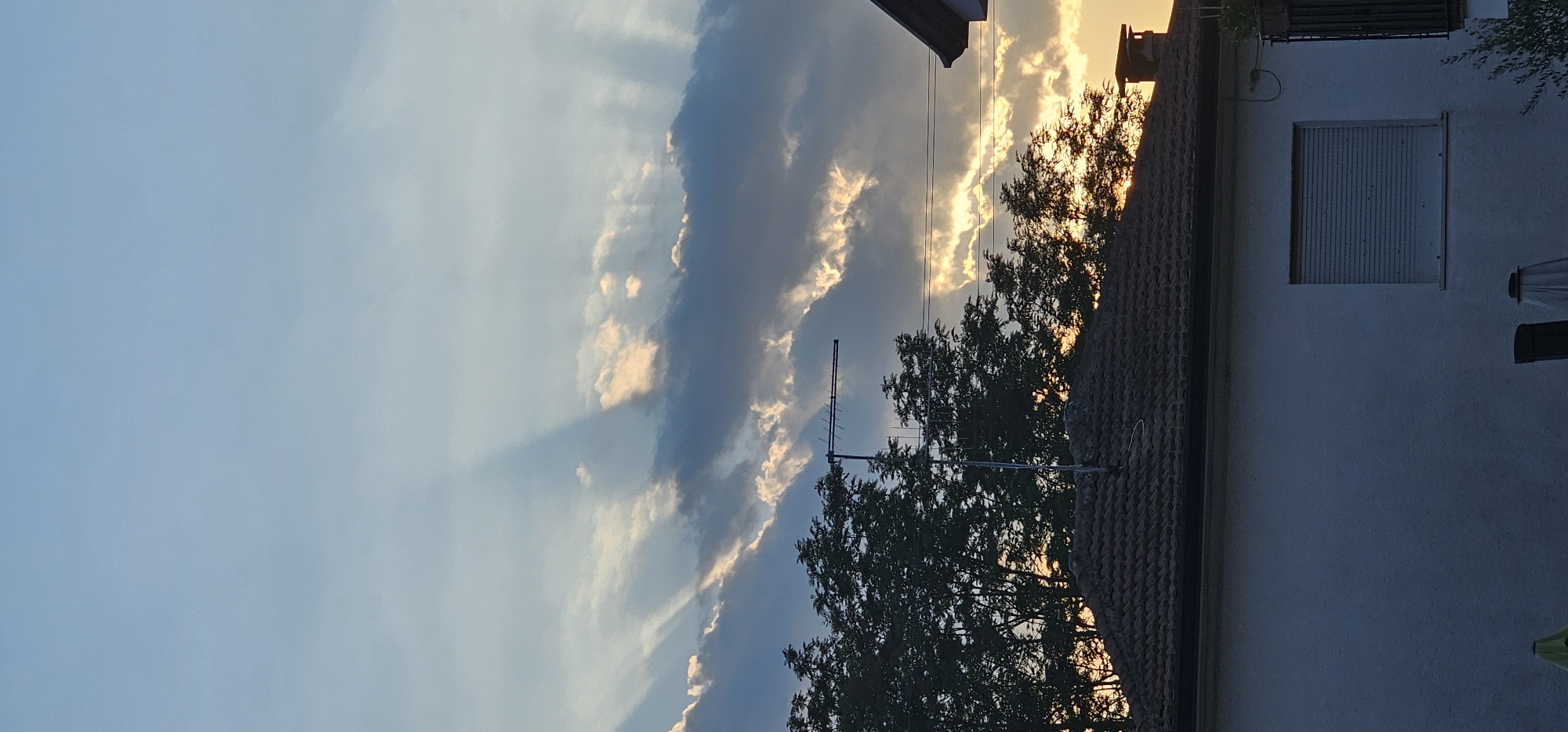 The image captures a sunset scene over a residential area in Villa Latina, Italy. The sky is a blend of soft blues and whites, with dramatic light rays piercing through scattered, dark clouds, illuminating patches of golden light. Silhouetted against the sky are the tops of lush green trees, and a television antenna. In the foreground, the tiled roof of a house is visible, with a white wall and a window covered by a closed, slatted blind. To the right, a portion of another house's roof and wall is visible, along with a chimney. The overall impression is one of a quiet, end-of-day atmosphere in a suburban setting.