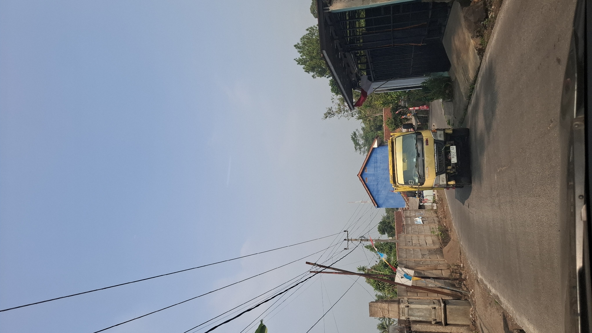 This is a photograph taken from a vehicle, looking forward down a paved road in Sirnagalih, Indonesia. The road curves slightly to the right. On the left side of the road, there is a concrete wall, a large concrete pillar structure, and further back, a single-story blue building with a red roof. Multiple electrical wires crisscross the sky above the scene.
In the center of the road, approaching the viewer, is a bright yellow Mitsubishi Fuso truck. The truck has a license plate that reads "F 8023 GS". Behind the truck, more buildings and trees are visible. On the right side of the road, there is a dark metal gate with a sloped roof structure extending from it. The sky is clear and blue with a few faint wispy clouds. The overall impression is a sunny day in a semi-urban or rural Indonesian setting.