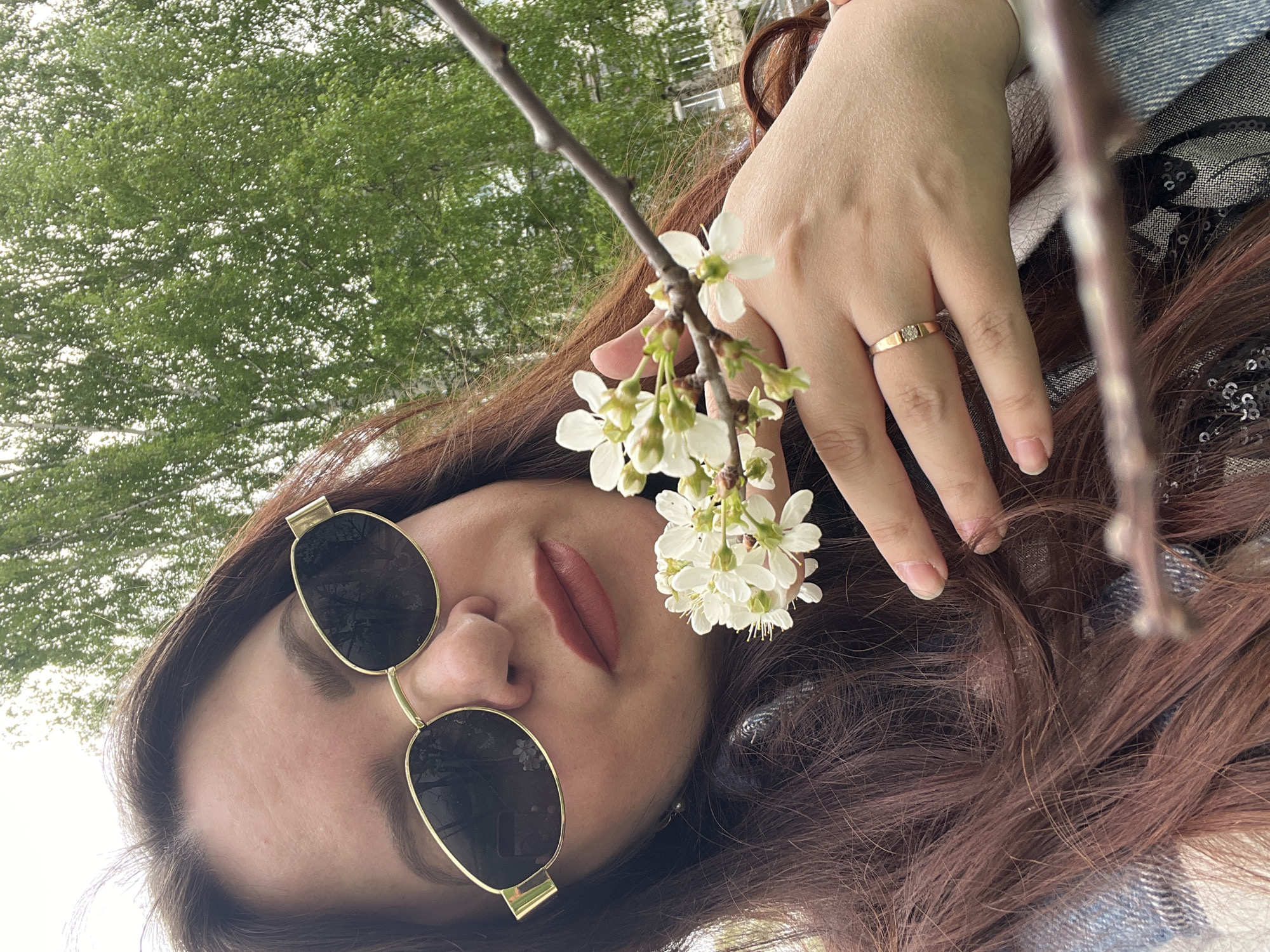 A woman lies on her back outdoors, presumably in Noviy, Russia, wearing gold-rimmed sunglasses. She holds a branch of white blossoms in her hand. She has brown hair, and her lips are painted a dark, matte color. A gold ring is visible on her ring finger. A tree canopy is visible in the background, along with the edge of a building.