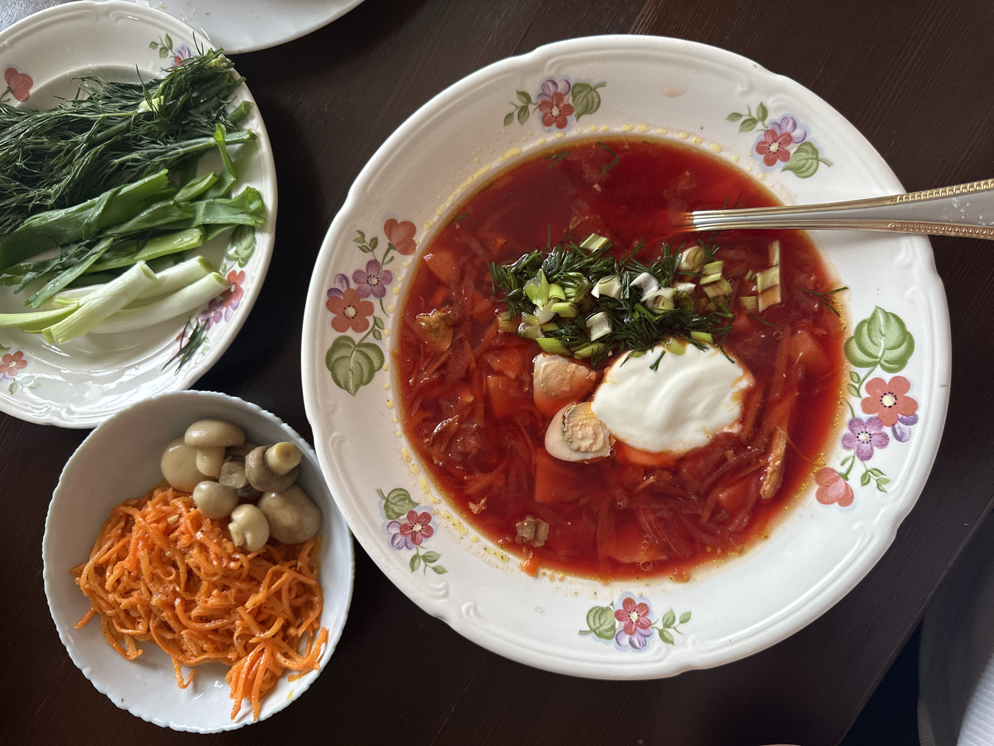 A tabletop scene shows a serving of borscht in a white floral-patterned bowl, garnished with sour cream, chopped green onions, and dill. Beside it are two smaller bowls. One bowl contains chopped carrots and mushrooms, while the other contains green onions and dill. A silver spoon rests in the borscht bowl. The setting is a dark wooden table. Based on the food composition, this image potentially reflects the cuisine of Chaikovsky, Russia.