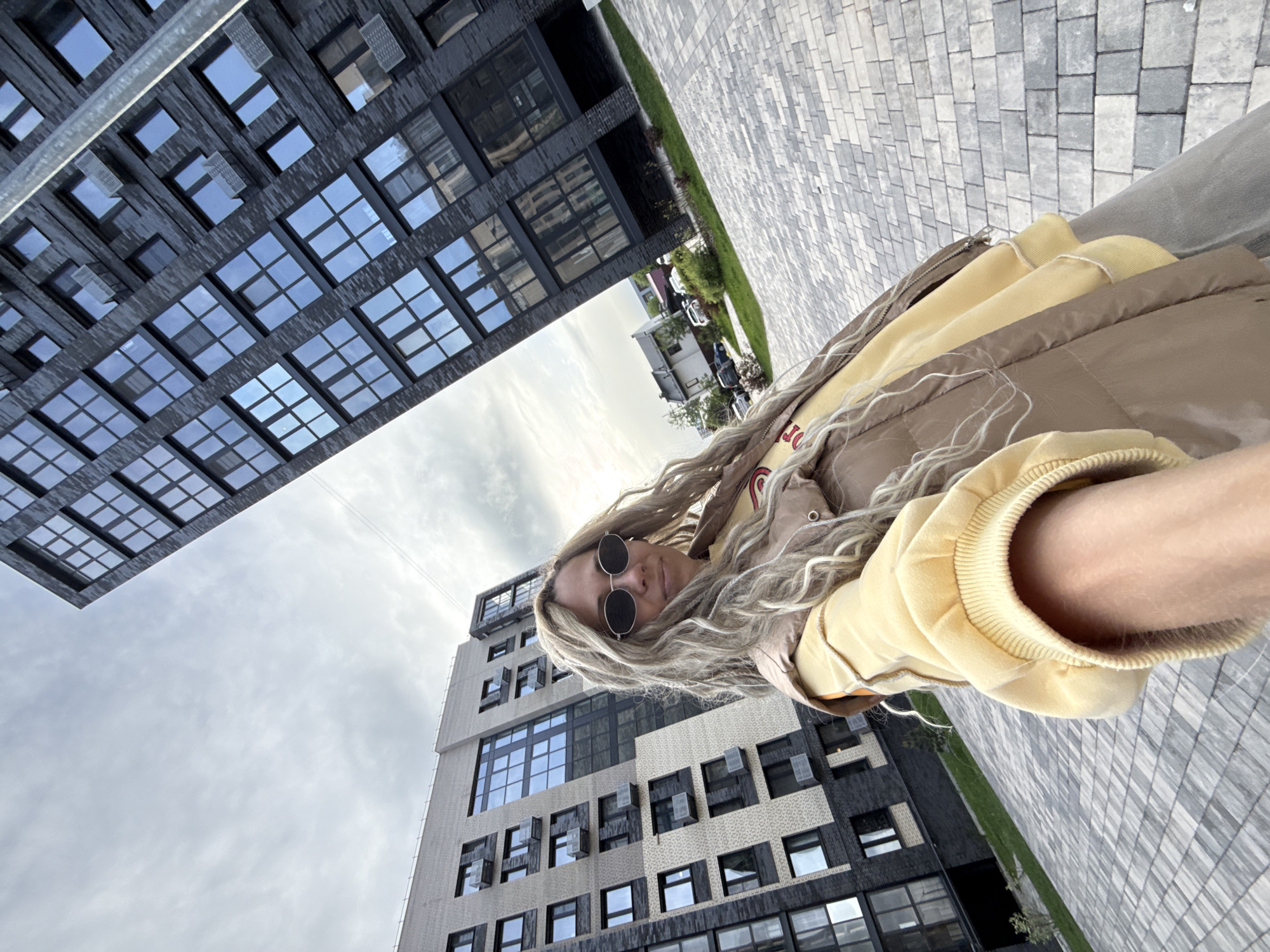A low-angle shot captures a woman wearing sunglasses, a yellow hoodie, and a brown jacket. She is surrounded by modern architecture, with tall buildings featuring grid-like windows visible in the background. The sky is overcast. The ground is paved with grey rectangular stones.