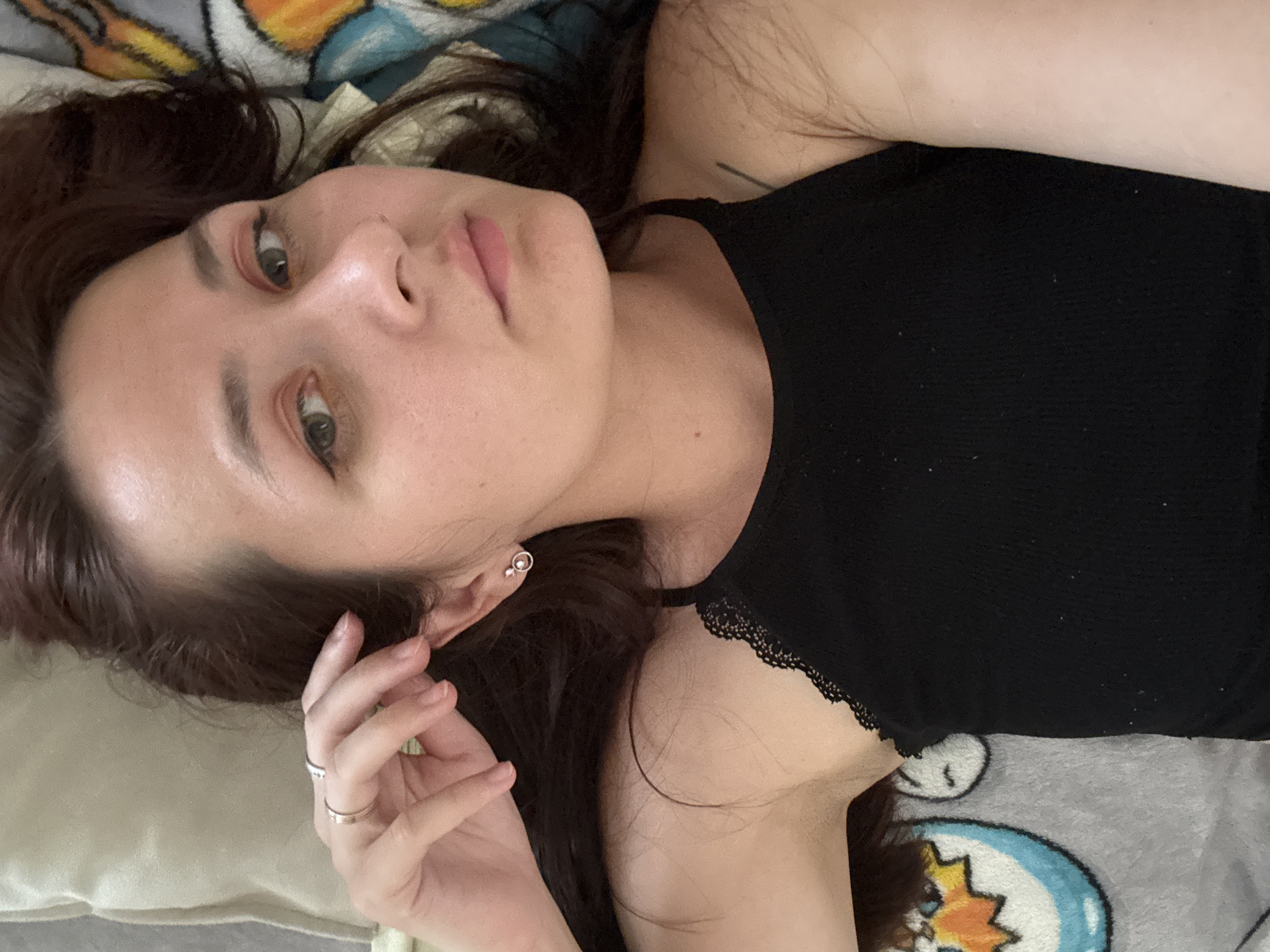A medium shot shows a young woman with dark hair lying on her back on a bed. She is wearing a black tank top with lace trim. Her head is slightly tilted to her right, and her left hand is raised, with her fingers touching her ear and temple. She is wearing two silver rings on her left ring finger and a small silver stud earring in her left ear. Her eyes are open, and she is looking towards the camera with a neutral expression. Her makeup includes eyeliner and pink eyeshadow. The background consists of a cream-colored pillow and a blanket with cartoon characters. The setting is an indoor environment, likely a bedroom. There is no information in the image that would allow for its location to be identified as Naberezhnye Chelny, Russia.