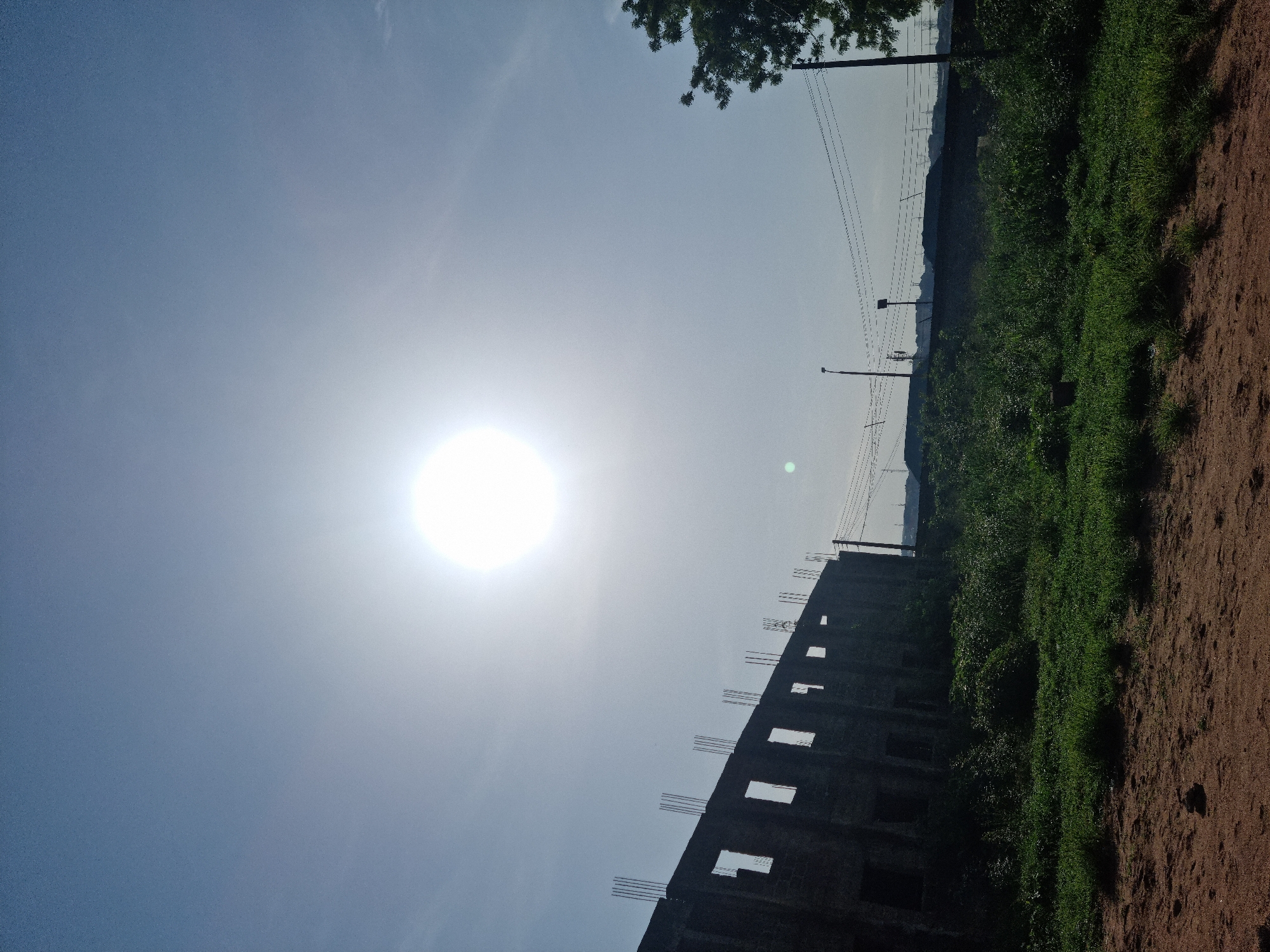 The image displays a bright, sunny daytime scene in what appears to be a developing area. The sun is intensely radiant in the upper left of the clear, light blue sky, casting a strong glare across the upper half of the frame. In the lower portion, a partially constructed concrete building stands, characterized by its unfinished state and exposed rebar protruding upwards from its top edge. Adjacent to the building, a vibrant strip of green grass and vegetation runs vertically, separating the structure from a broad expanse of reddish-brown dirt ground on the far right. Several utility poles with numerous power lines are visible in the background, extending vertically. A tree branch with green foliage peeks into the frame from the upper right corner. The strong sunlight and the nature of the construction, combined with the utility infrastructure and natural elements, suggest a warm, daytime setting in a transitioning landscape, consistent with the provided location context of Onitsha, Nigeria. There are no people visible, and no specific activity is taking place beyond the depicted environment. No visible text is present.
