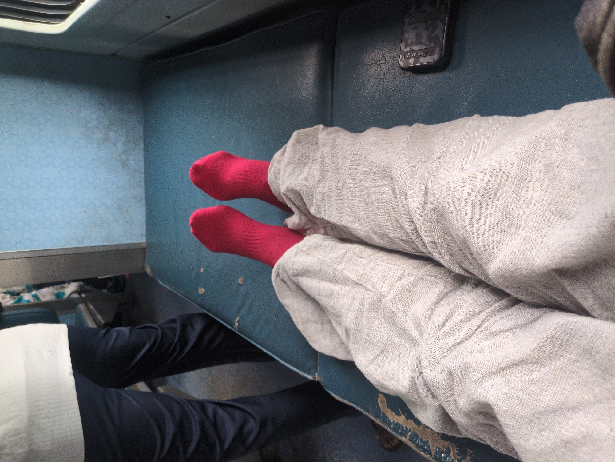 The image depicts a person's legs and feet, clad in light-colored pants and bright red socks, resting on a blue seat. The seat appears to be part of a train compartment, indicated by the presence of a blue patterned wall and a window frame in the background. A mobile phone with a camouflage pattern is visible on the right side of the seat. To the left, another person's dark pants and white shirt are visible, suggesting a shared space. The scene is casual and relaxed, indicative of travel. Given the context of travel, this scene could be taking place on a train heading towards or departing from Jhandipur, India, a small town known for its railway station.