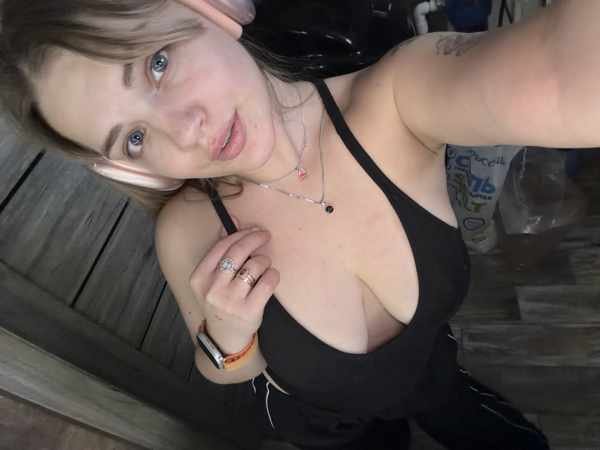A self-portrait features a young woman indoors in Большая Шильна, Russia, captured from a high angle. She has light brown hair framing her face, light skin, and prominent blue eyes that look directly at the camera with a slight smile.
She is wearing a black sports bra or tank top, revealing her chest, and dark pants with a white stripe visible on one leg. On her left hand, she wears several rings, including one with a large oval-shaped stone and another with a smaller, possibly red, stone. Her left wrist is adorned with a silver-toned smartwatch featuring an orange band. Around her neck, two necklaces are visible: one with a small red, rectangular charm (possibly a stylized milk carton or brick shape) and another with a small, dark, circular pendant. A tattoo is partially visible on her upper left arm.
The background on the left side of the image consists of wooden planks, suggesting a floor or wall. On the right, the background is darker and more cluttered, showing what appears to be white pipes and a large grey bag. Visible Cyrillic text on the bag includes "СОЛЬ" (Salt), "БАННАЯ" (Bath), and "ГОСТ" (GOST, a Russian quality standard), indicating it is likely a bag of bath salts. The lighting is soft, highlighting her features. The image conveys a casual, close-up moment.