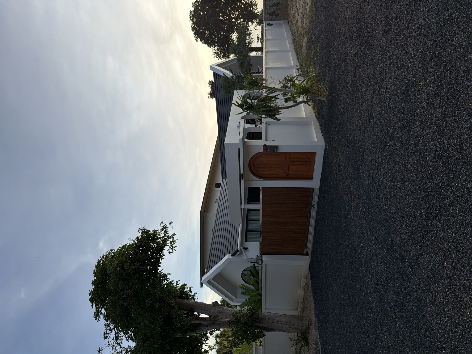 This is a landscape-oriented, low-angle, outdoor photograph of a modern white house with dark gray tile roofing and wooden gates. The house has a contemporary design with sharp angles and a prominent triangular peak over an arched entrance. Lush green trees are visible in the foreground and background. The sky is overcast with patches of sunlight, suggesting it is either early morning or late afternoon. The ground is covered in dark gravel, and a white wall surrounds the property. There is no visible text in the image. The scene appears to be a residential property in Baan Plai Laem, Thailand. The overall mood is serene and peaceful, with the natural elements of trees and sky complementing the modern architecture.