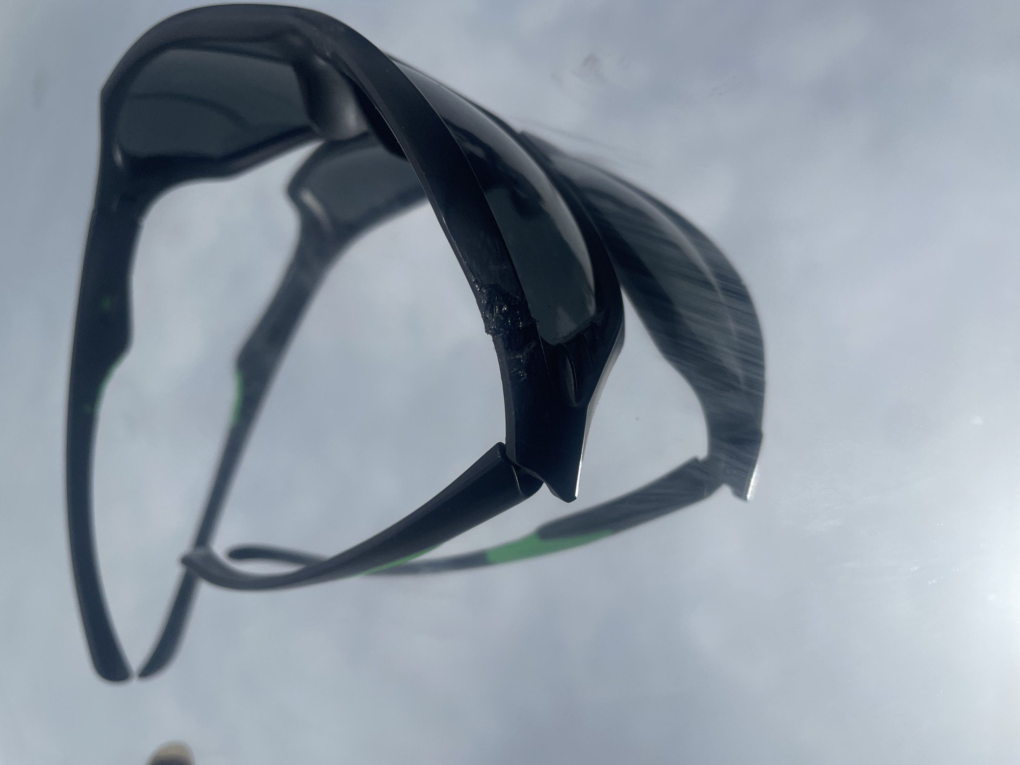 A close-up, low-angle view of a pair of black sunglasses with green accents, seemingly resting on a reflective surface against a cloudy, bright sky. A portion of a light-colored object is visible in the bottom left corner. The sunglasses appear to be slightly damaged on the left side of the frame.
There is no information to suggest that the image was taken in Jos, Nigeria.