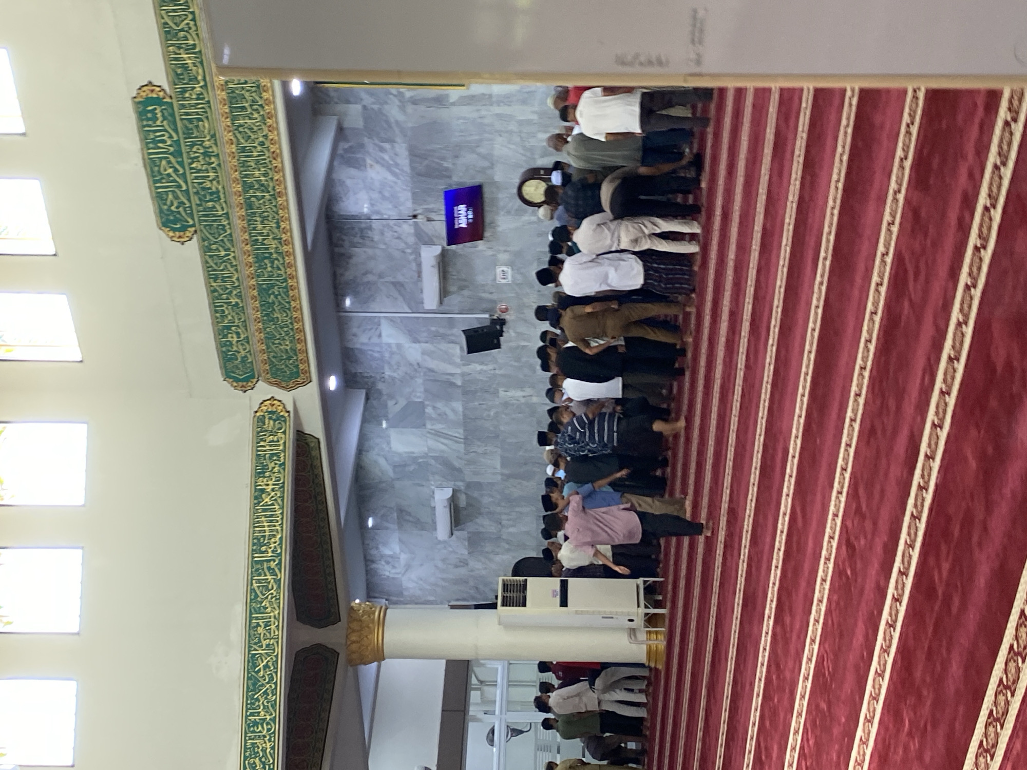 This image captures the interior of the Islamic Center Masjid Agung in Lhokseumawe, Indonesia, likely during prayer time. A congregation of men, dressed in various attire including sarongs and casual clothing, stand on a red carpet patterned with white and gold stripes, facing the prayer direction. Some are in the process of praying, indicated by their postures. The mosque features ornate green and gold Islamic calligraphy along its walls and ceiling. A large white pillar with gold accents supports the structure, and a tall white air conditioning unit stands nearby. In the background, a large clock is visible, and a digital display shows "ASHAR" with prayer timings. The walls are adorned with grey marble-like tiles. The overall scene depicts a communal religious gathering within a grand and well-decorated mosque.