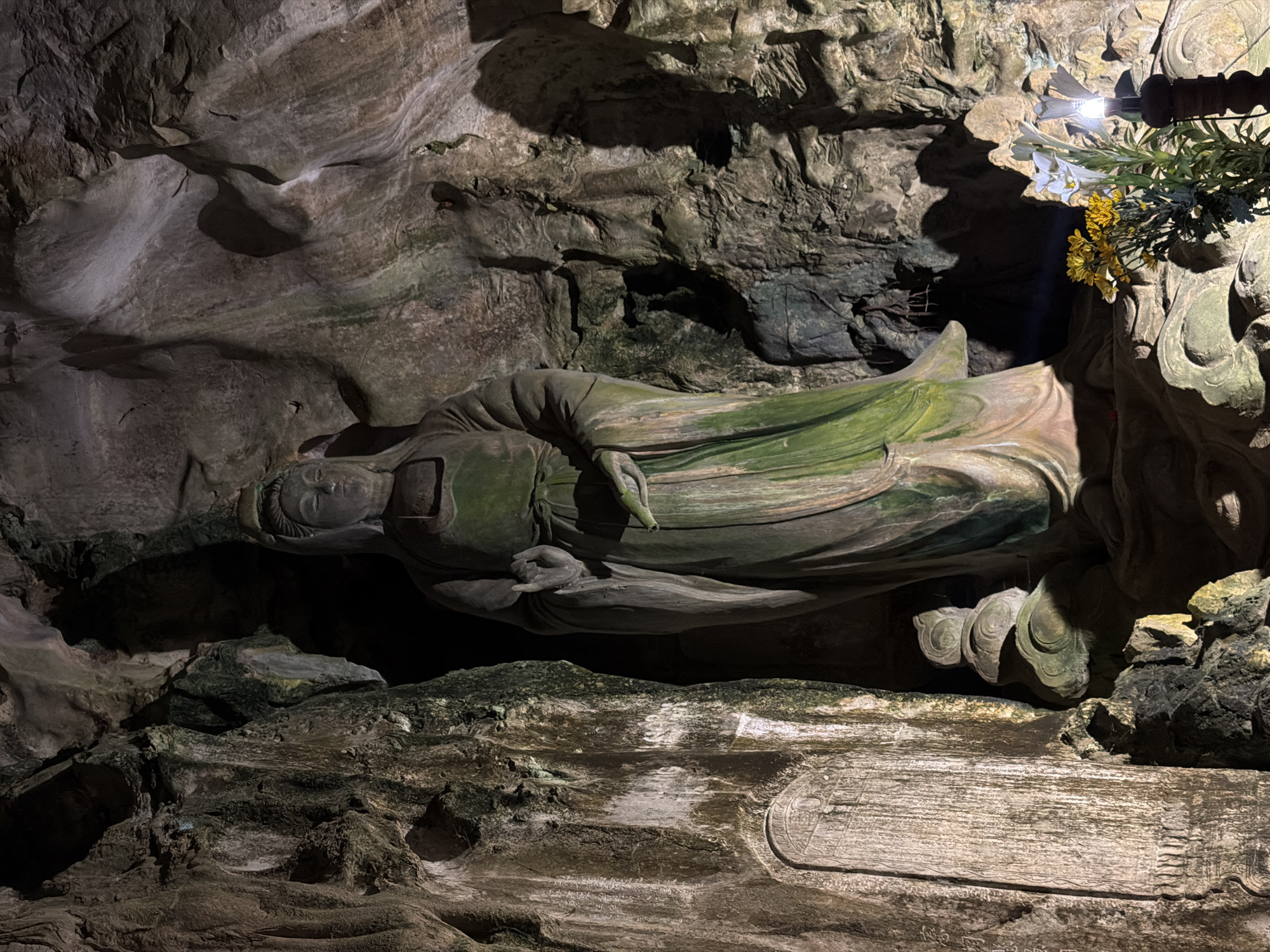 This vertical image presents a large, reclining Buddha statue nestled within a natural cave environment in Đà Nẵng, Vietnam. The central subject is a serene statue, likely carved from stone or wood, depicted lying on its right side with its head resting on a raised platform or pillow. The figure is draped in robes that exhibit a greenish, moss-like patina, suggesting age or the cave's humid conditions. Its left hand is gently positioned near its chest.
The setting is a rugged, dimly lit cave with irregular rock formations forming the walls and ceiling. These natural surfaces display varying textures and colors, including shades of grey, brown, and areas of green mineral or moss growth. Strategic artificial lighting, originating from the upper right, illuminates the Buddha's face and upper torso, casting pronounced shadows that enhance the depth and texture of the cave and the statue. Adjacent to the Buddha's head, a small arrangement of artificial white lilies and yellow chrysanthemums is visible, also highlighted by the light.
Below the reclining Buddha, a flat, carved stone base is evident. Faint, etched characters or symbols, possibly an Asian script, are visible along this surface. Towards the right side of this base, a distinct circular or oval carving with intricate patterns or additional text is present, though not clearly legible. The scene evokes a sense of spiritual reverence and ancient tranquility within the natural grotto.