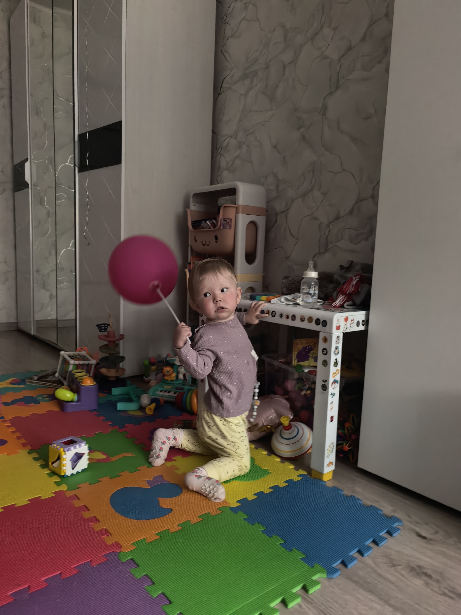A young child, likely a toddler, is the central subject of the image, kneeling on a colorful foam play mat indoors. The child has light brown hair and a fair complexion, wearing a light purple long-sleeved top and yellow patterned pants with white socks. They are holding a bright pink balloon attached to a stick, looking thoughtfully towards the viewer's left with a neutral to slightly curious expression.
The setting appears to be a child's play area within a home in Sterlitamak, Russia. The floor is largely covered by a multi-colored interlocking foam puzzle mat, featuring various hues like red, orange, yellow, green, blue, and purple. One piece has a blue duck-shaped cutout. Numerous toys are scattered around the mat, including a white cube-shaped toy, a purple and yellow ball-drop toy, a toy piano/keyboard, and other small play items.
In the background, a white shelving unit or table is visible to the child's right, holding a baby bottle, a pink plastic storage caddy, and other objects. The walls feature a light-colored wallpaper with a marbled or textured pattern. To the left, a large wardrobe with mirrored panels and white frames reflects parts of the room. A plain white cupboard or wall unit stands on the right side of the image. The lighting is somewhat dim, suggesting an indoor shot without intense natural light. No visible text is legible in the image.