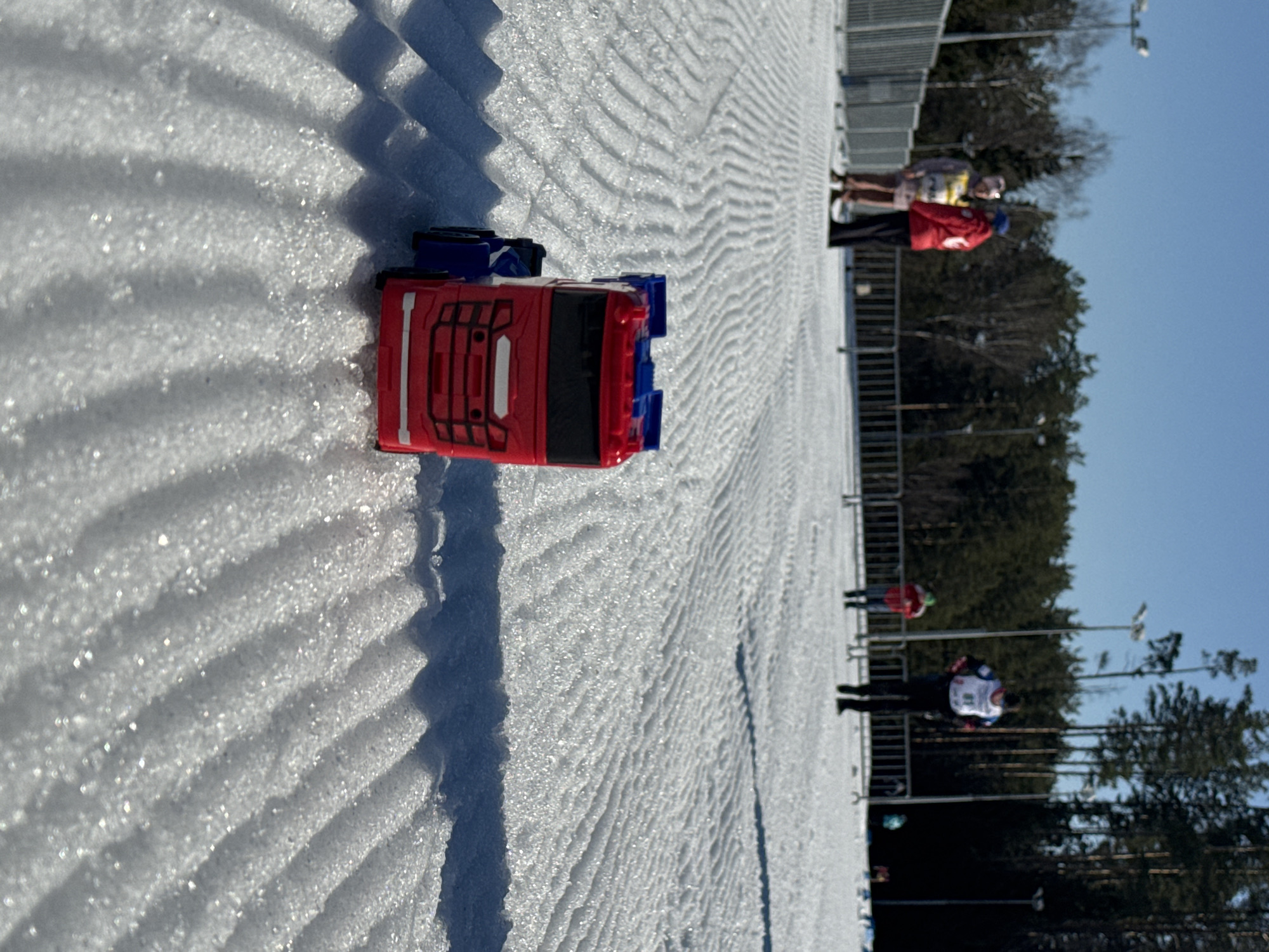 A red and blue toy semi-truck is positioned on a snowy, textured surface. The texture appears to be from a ski trail. In the background are several people standing near a fence line, and beyond the fence line are trees and a clear, blue sky. The location is Прикамский, Russia.