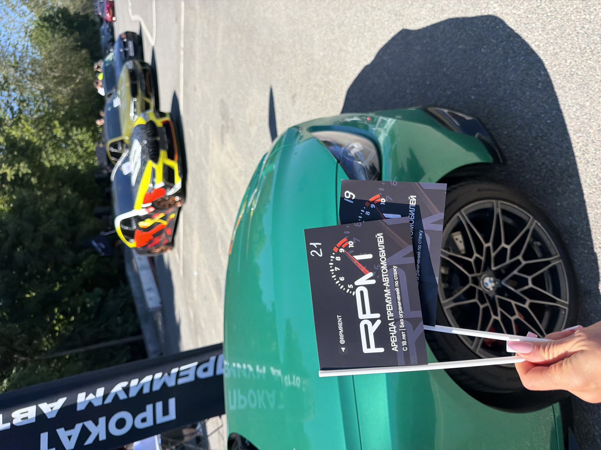 In Saint Petersburg, Russia, on a sunny day, a woman's hand with pink nail polish is holding two small flags in front of a green BMW car. The flags are branded with "RPM" and feature a tachometer graphic, indicating car rental services. Behind the green BMW, several other cars are parked in a line on an asphalt surface. In the background, lush green trees line the edge of the parking area. A large black banner with white lettering, partially visible on the left side of the image, also advertises "PROKAT PREMIUM AVTO," suggesting a premium car rental service. The overall scene appears to be an outdoor event or exhibition related to car rentals.