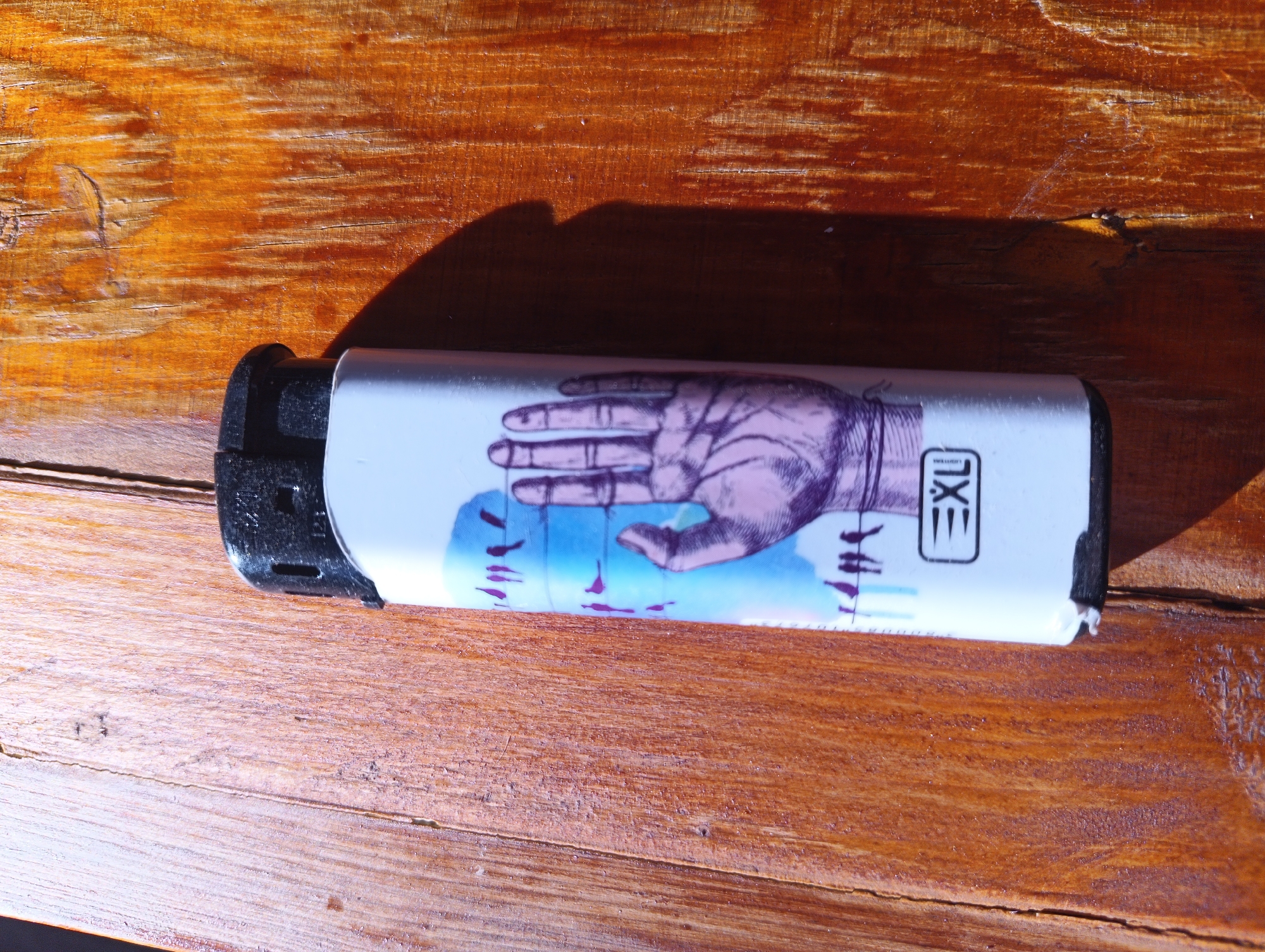 A cigarette lighter lies on a wooden surface. The lighter has a white casing with a graphic design of a hand holding strings that control small figures. A black brand logo, appearing as "EXU" in a rectangular box, is visible on the right side of the casing. The wooden surface is made of planks and exhibits a reddish-brown color with visible grain and some wear. A shadow is cast on the surface to the left of the lighter, suggesting a light source from the right. The scene is indoors, with no discernible urban elements to place it specifically in Karnobat, Bulgaria.