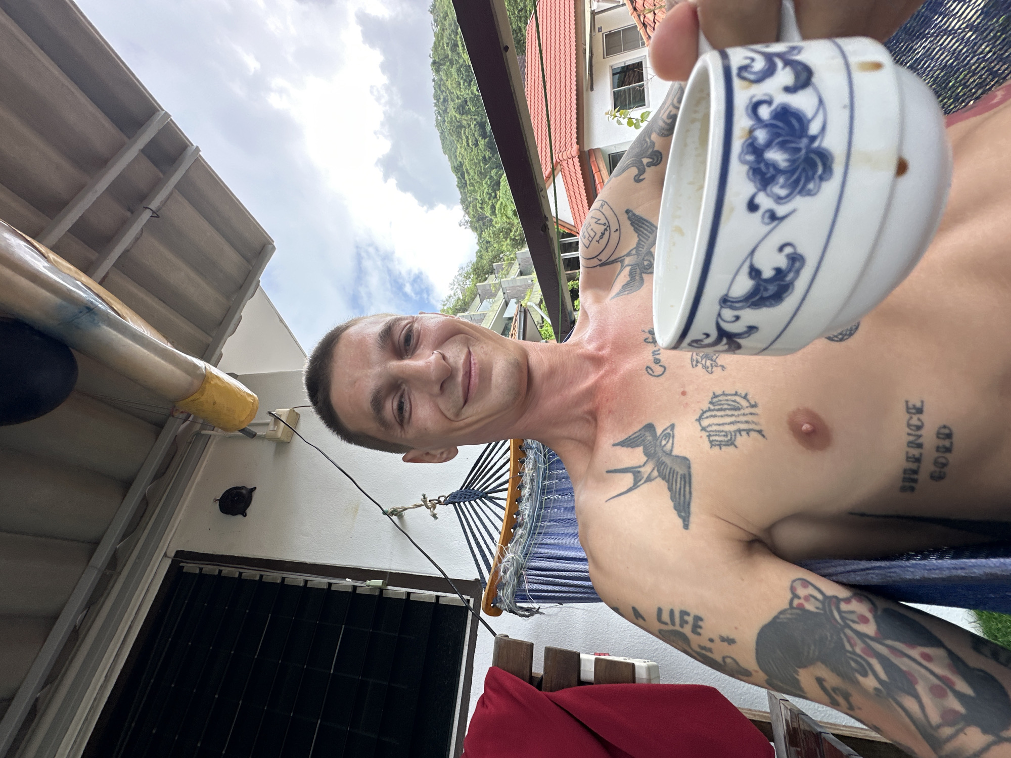 In Karon, Thailand, a shirtless man with numerous tattoos, including swallows and text, smiles at the camera while holding a cup of coffee. He is on a balcony, with a hammock and a red cloth visible behind him. In the background, buildings with red tiled roofs and lush green hills are present under a cloudy sky.