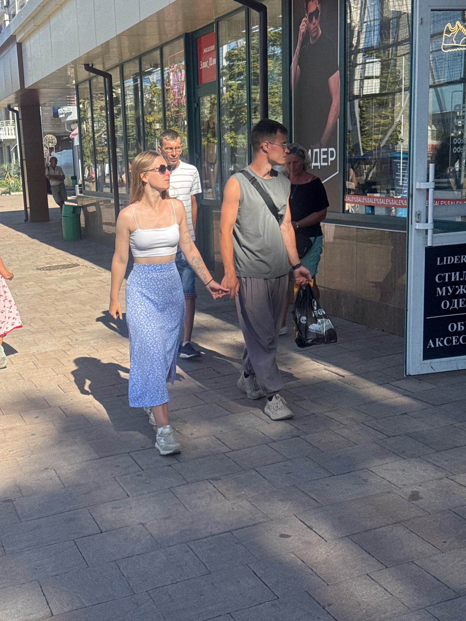 In Anapa, Russia, a couple, a man and a woman, walks hand-in-hand on a sunlit pedestrian street lined with shops. The woman, on the left, wears a white crop top and a long blue skirt with a floral pattern. The man, on the right, is dressed in a gray sleeveless shirt and gray sweatpants. He carries a black shopping bag. Behind them, other pedestrians are visible, including a man in a striped shirt and glasses, and a woman in a black top. The storefronts display advertisements, one featuring a large image of a man in sunglasses. The ground is paved with rectangular stone tiles, and shadows are cast by the buildings and people.