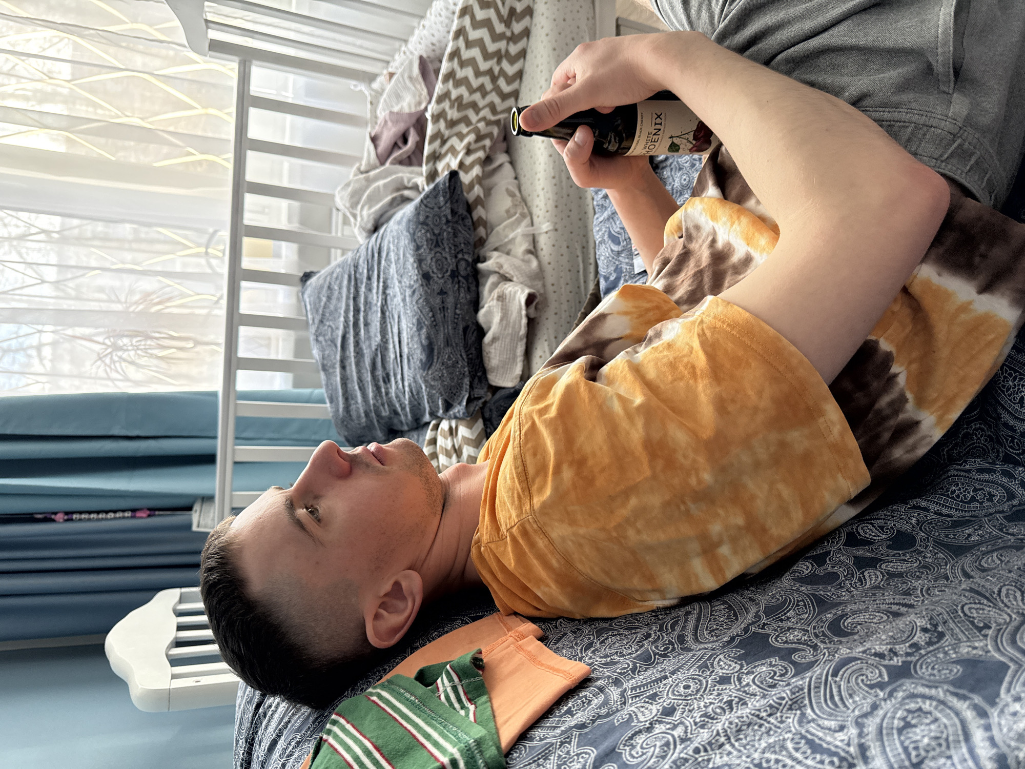 A man is lying on a bed holding a bottle labeled "WHITE PHOENIX MINERAL BLACK CHERRY". He is wearing a tie-dye t-shirt and grey pants. A green and orange striped garment is near his head on the bed. The room includes a crib and a window with closed blinds. The exact location in Moscow, Russia, cannot be determined from this image.