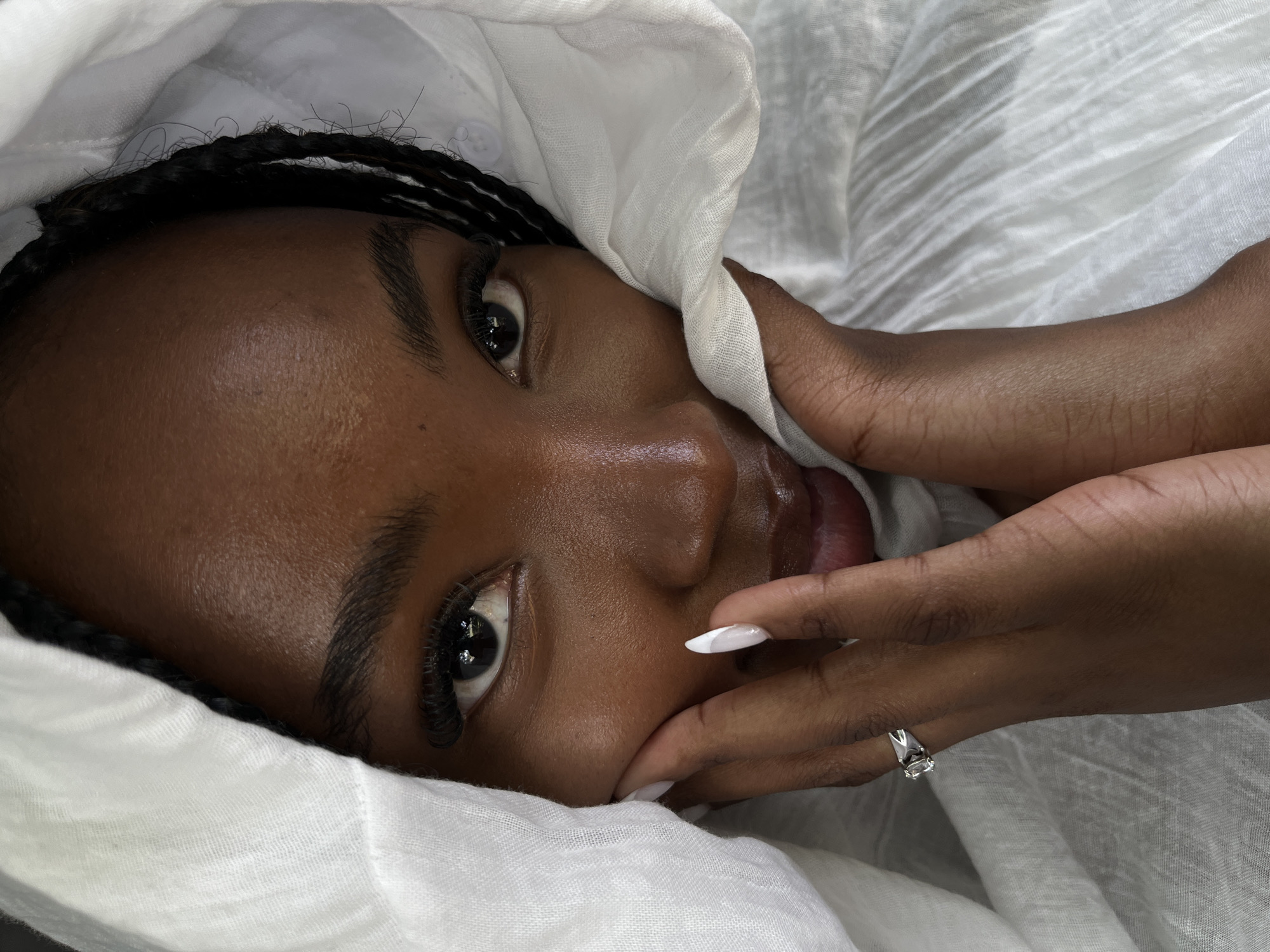 A close-up, head-and-shoulders portrait shows a young Black woman partially shrouded in white fabric. Her dark braids are visible peeking out from under the fabric. She is looking directly at the camera with her eyes open, and her hands are placed on either side of her face, with her fingers gently touching her cheeks. Her fingernails are painted white with a French tip. A diamond ring is visible on her left ring finger. The background consists of more white fabric. The lighting suggests a soft, natural light source. The image is a portrait taken from a close perspective, focusing on the subject's face and hands. There is no indication of the location being Kazan, Russia.