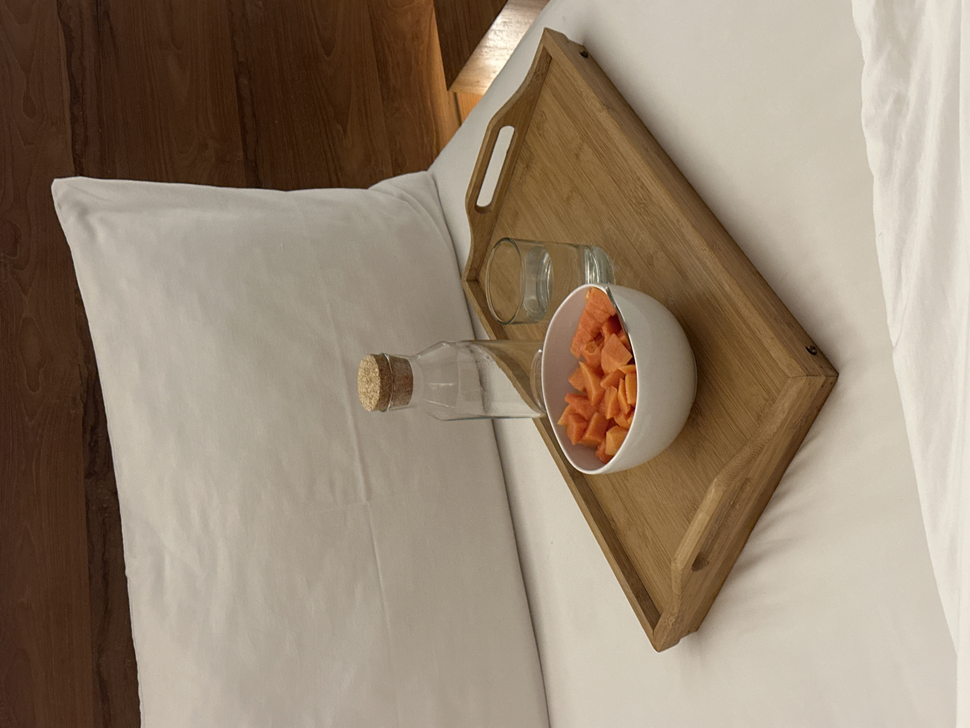 The image shows a bed tray with a bowl of papaya, a glass, and a bottle of water. The tray sits on a white bedsheet. The background features a wooden headboard. The image was captured indoors in Sukawati, Indonesia.