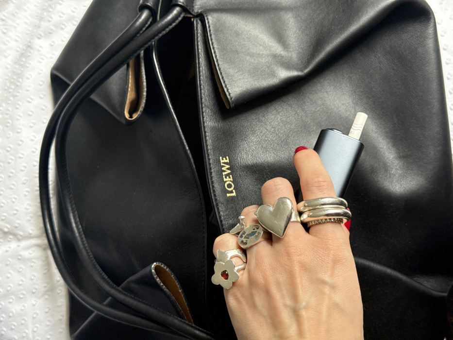 A close-up, overhead view depicts a woman's hand holding a heated tobacco device over a black leather tote bag in London, United Kingdom. The bag, identifiable by a gold 'LOEWE' logo on an interior panel, is partially open, revealing a lighter-colored interior lining. The hand, with red-painted fingernails, wears multiple silver-toned rings: a flower-shaped ring with a red central gem on the index finger, a large heart-shaped ring on the middle finger, and three bands on the ring finger (two plain, one thin with dark stones). The hand securely grasps a dark grey, rectangular heated tobacco device with a white filter-like stick protruding from one end. The background consists of a white, textured fabric surface.