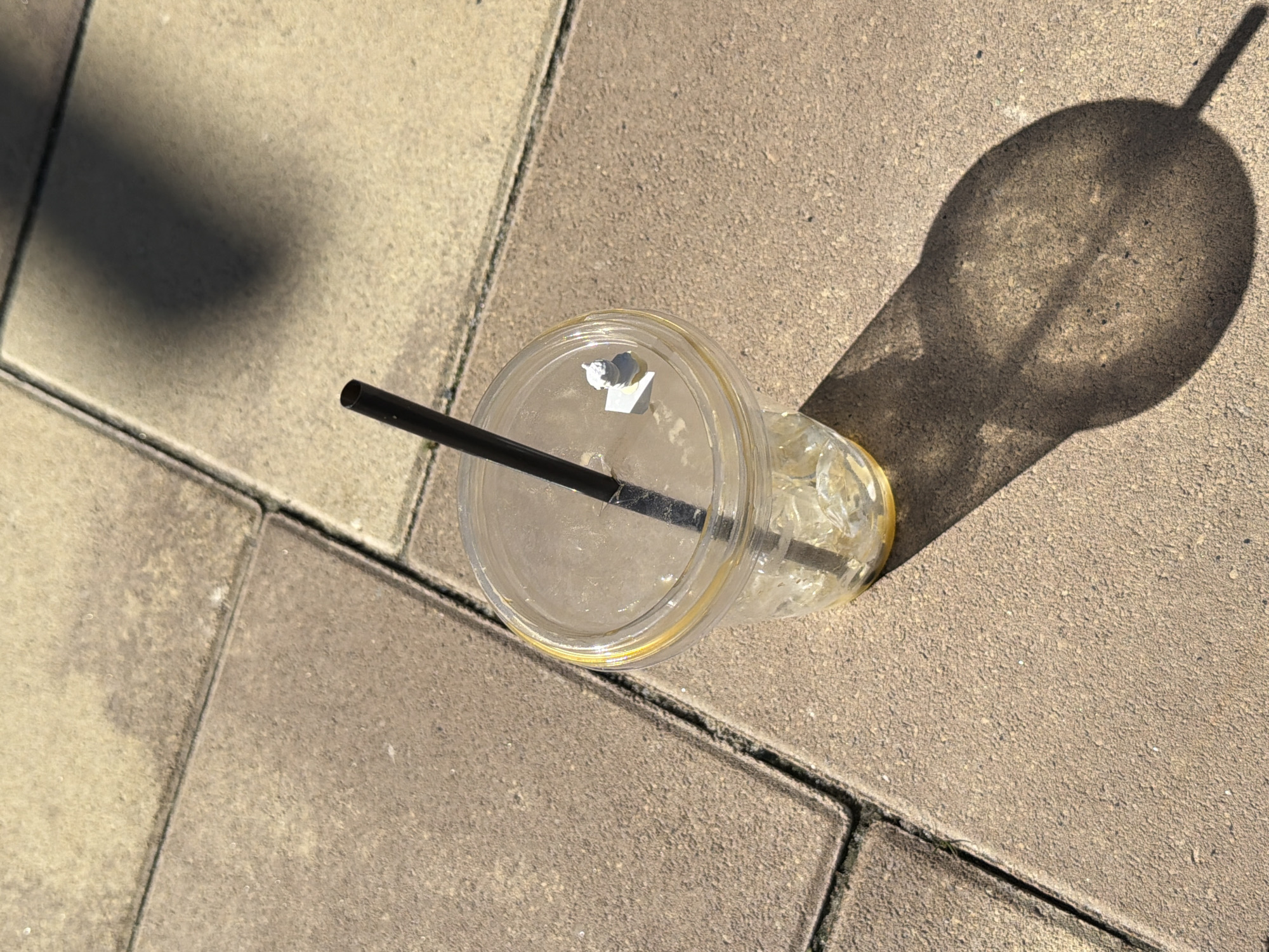 An empty plastic cup with a black straw sits on a paved surface. The cup is clear and contains ice cubes and a small amount of liquid at the bottom. There is a small white object on top of the lid of the cup, next to the straw. The paved surface is made of rectangular tiles, some of which are lighter in color than others. A shadow of the cup and straw is cast on the ground to the right. The scene is outdoors, likely in a public area. The image does not contain any specific identifying features that would allow for the determination of its location in Kyiv, Ukraine.