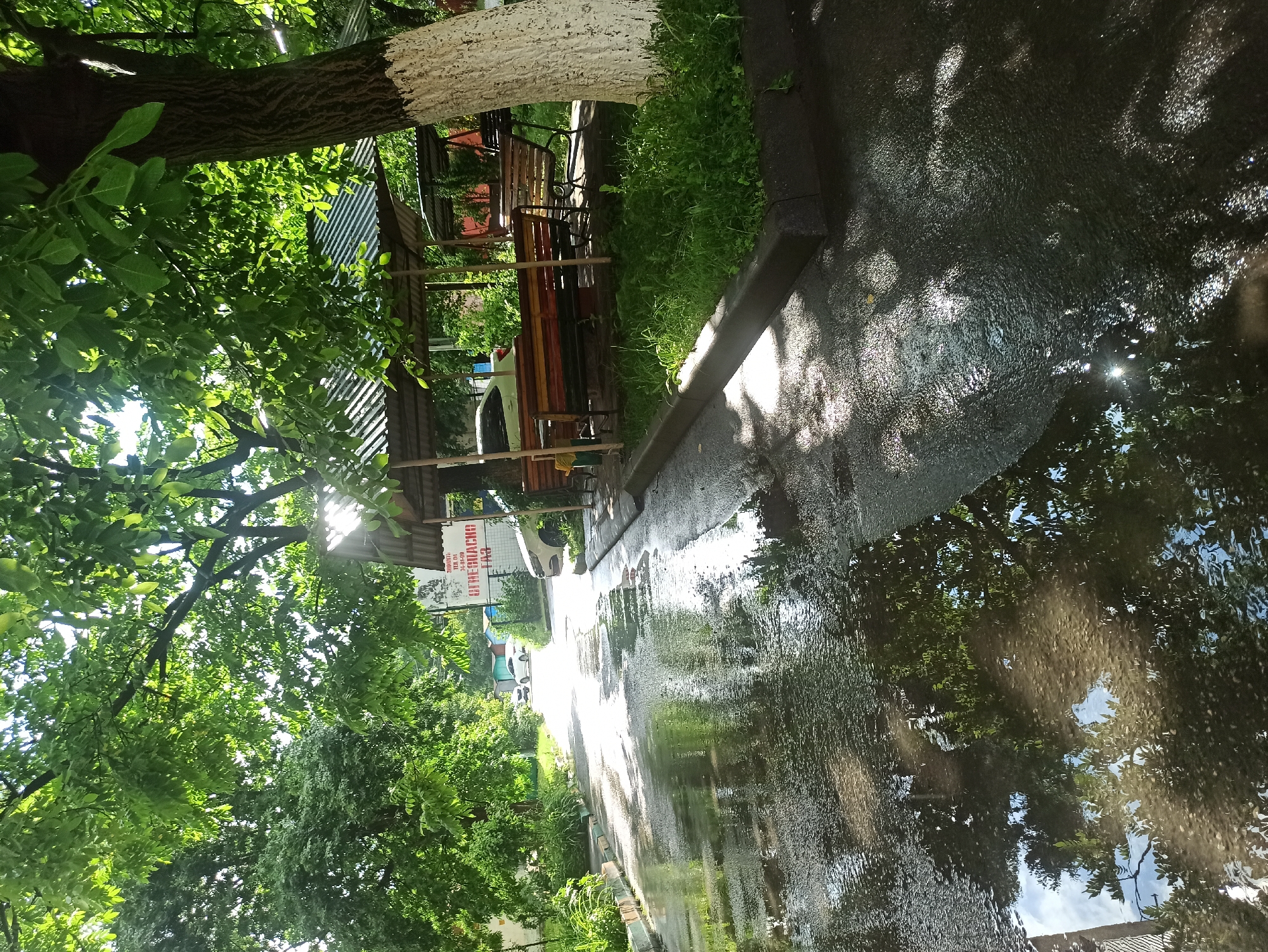 The image depicts a wet asphalt road reflecting sunlight and trees. On the right side of the road, there's a covered seating area with benches and a table, set against a backdrop of trees and a building. The building has a sign with Russian text. There are trees with green foliage throughout the scene. The road appears to be in a residential or communal area, possibly a courtyard or park. The reflection in the water on the road shows the sky, trees, and some buildings. There are no people visible in the image. The overall impression is one of a quiet, possibly post-rain, outdoor scene in a suburban or urban environment. The location is identified as Atabe, Georgia, though the visual elements themselves do not provide definitive clues to this specific location.