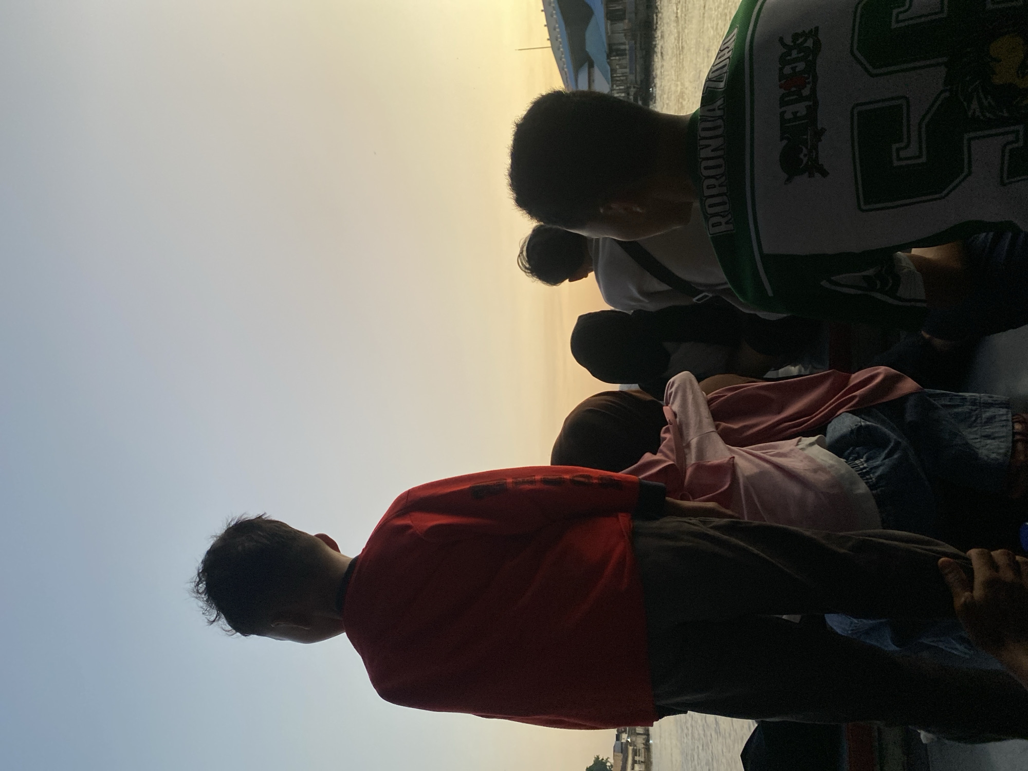 The image depicts a group of people, silhouetted against a sunset sky, likely on a boat in Banjarmasin, Indonesia. The primary subject is a young boy in the foreground, facing away from the camera, wearing a red long-sleeved shirt and dark pants. He appears to be standing, with another person's arm supporting him from behind. Behind him, several other individuals are visible, including a person in a pink top and blue jeans, and others wearing dark clothing. Further in the background, a person in a green and white shirt with "RORONOA ZORO" and "ONE PIECE" graphics is seated. The environment includes a body of water, likely a river, with buildings and structures on the opposite bank. The sky exhibits a gradient of colors, transitioning from light blue at the top to soft yellow and orange at the horizon, characteristic of a sunset. The overall scene suggests a casual outing or travel by boat during the late afternoon or early evening.
