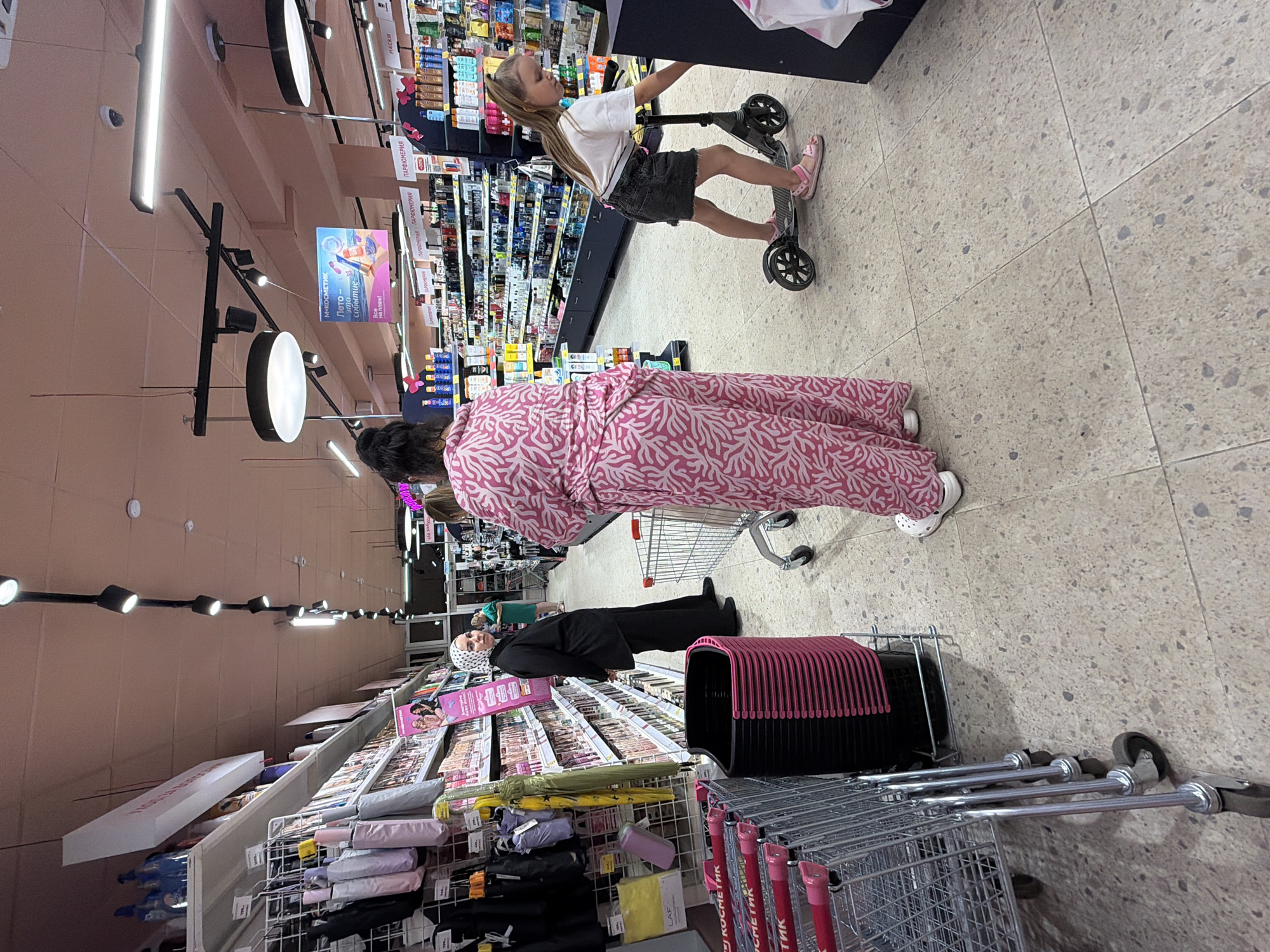 The image depicts the interior of a brightly lit supermarket aisle, likely located in Sochi, Russia, given the Russian signage visible. In the foreground, a row of shopping carts is parked on the left. Further into the aisle, a woman wearing a black headscarf and a matching black outfit stands looking towards a shelf. To her right, a person in a long, pink and white patterned robe is facing away from the camera. On the right side of the image, a young girl with blonde hair is riding a scooter. The shelves are stocked with various products, including what appear to be cosmetics and personal care items, with promotional posters displayed. The floor is tiled with a speckled, light-colored material. The overall atmosphere suggests a typical retail environment.