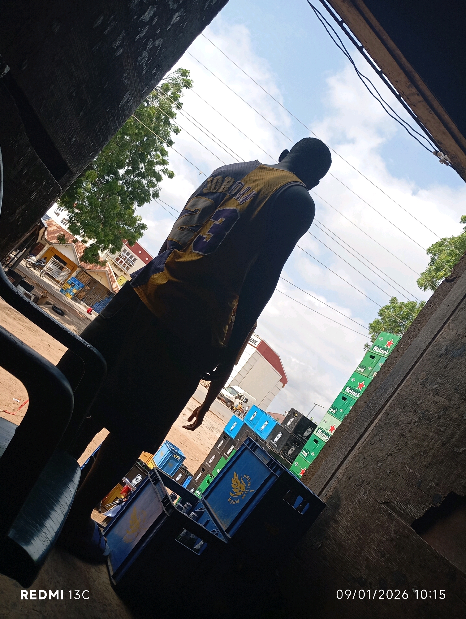 A low-angle outdoor medium shot captures a young Black male from behind, wearing a yellow and purple basketball jersey with the number 23. He stands facing away from the camera, looking towards buildings and trees. The scene is set in an urban environment, likely in Shendam, Nigeria, judging by the presence of Heineken crate stacks and local structures. The foreground is dominated by dark, textured surfaces, possibly a wooden overhang or structure, with part of a black chair visible on the left. In the mid-ground, stacks of blue and green plastic crates, some with "Heineken" branding, are arranged. A street with a white vehicle and a motorcycle is visible behind the crates. The sky above is partly cloudy. The bottom right corner displays the date "09/01/2026" and time "10:15" with "REDMI 13C" branding in the bottom left. The overall impression is a snapshot of daily life in a Nigerian city, possibly a market or a commercial area.
