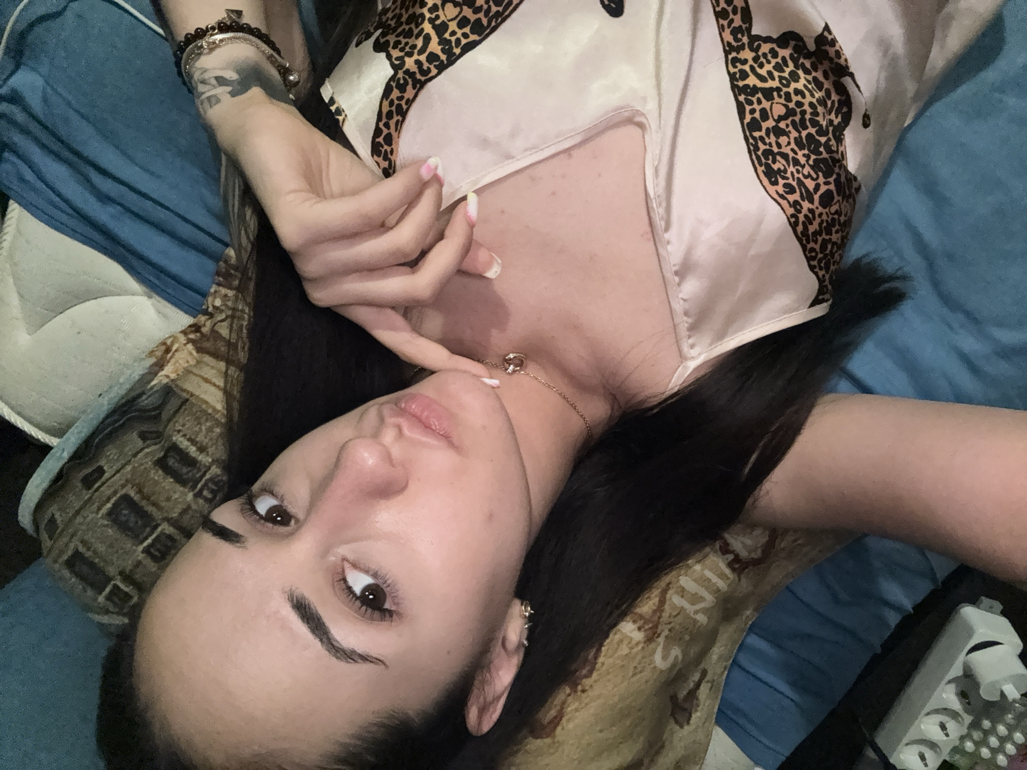 A woman with dark hair and brown eyes is lying down on a bed, looking at the camera. She is wearing a light-colored pajama top with a leopard print design. She has a necklace with a circular pendant and bracelets on her wrist. There is a tattoo visible on her forearm. The background shows a blue blanket and a patterned pillow. The lighting suggests an indoor setting, possibly a bedroom. This image does not provide any information about the location in the city of Shumen, Bulgaria.