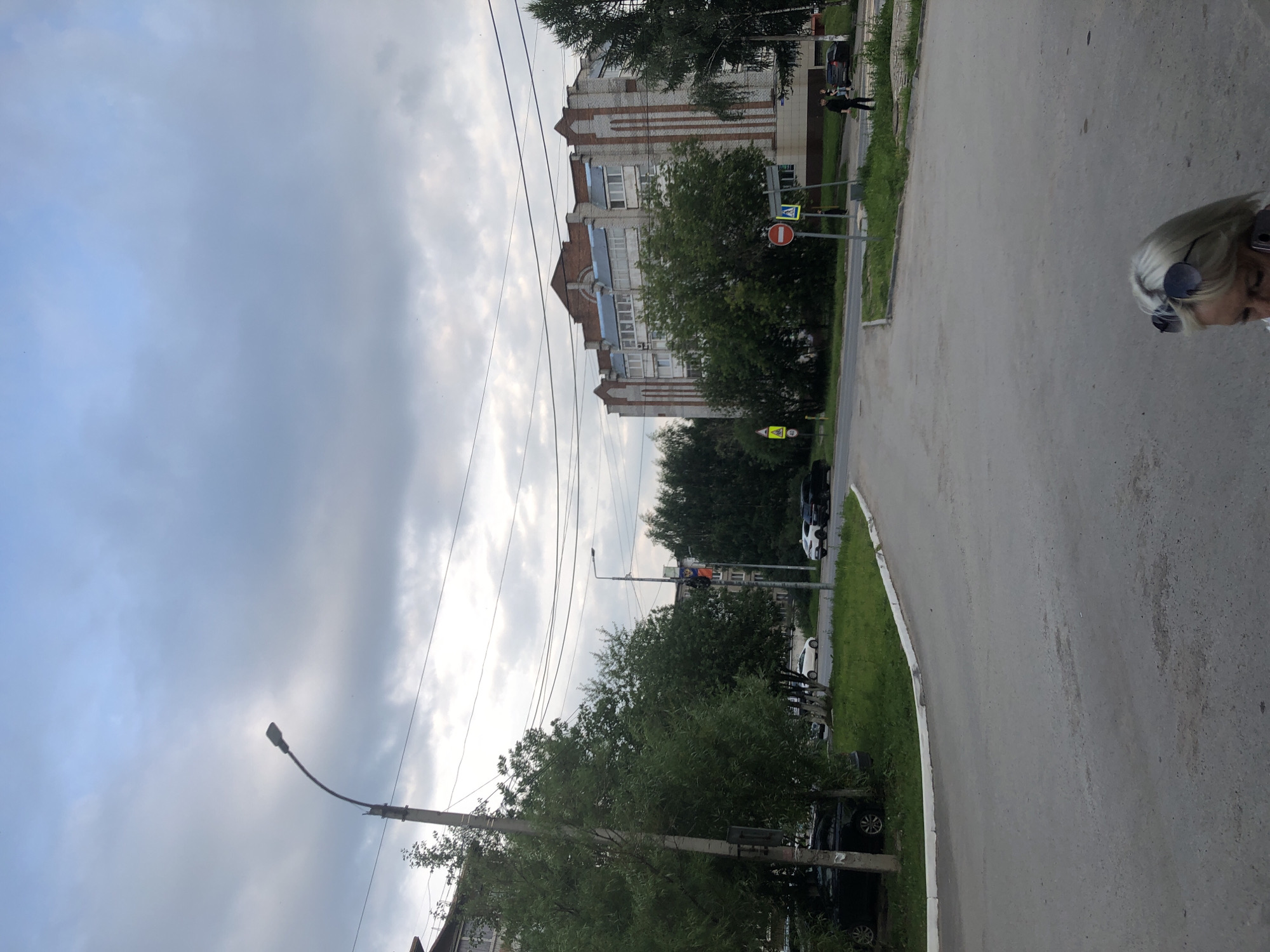This media file, taken in Solikamsk, Russia, depicts a street scene under a cloudy sky. The image is oriented vertically, with the sky occupying the top portion and the ground level at the bottom.
The foreground features a paved road and a grassy verge. On the right side of the frame, a person with blonde hair and sunglasses is partially visible, seemingly walking or standing on the pavement.
Further down the street, the scene includes several buildings, trees, and traffic signs. One building is a multi-story apartment block with brick and white facades, and balconies. Across the street from this building, there is a pedestrian crossing indicated by a crosswalk sign and a "no entry" sign. Trees are planted along the street, and there are several parked cars visible, including a black car and a white car. A lamppost is also present, extending into the sky.
The sky is filled with heavy, grey clouds, suggesting overcast weather, possibly indicating early morning or late afternoon given the soft light. The overall impression is that of a typical residential area on a cloudy day. There is no visible text that can be read clearly.