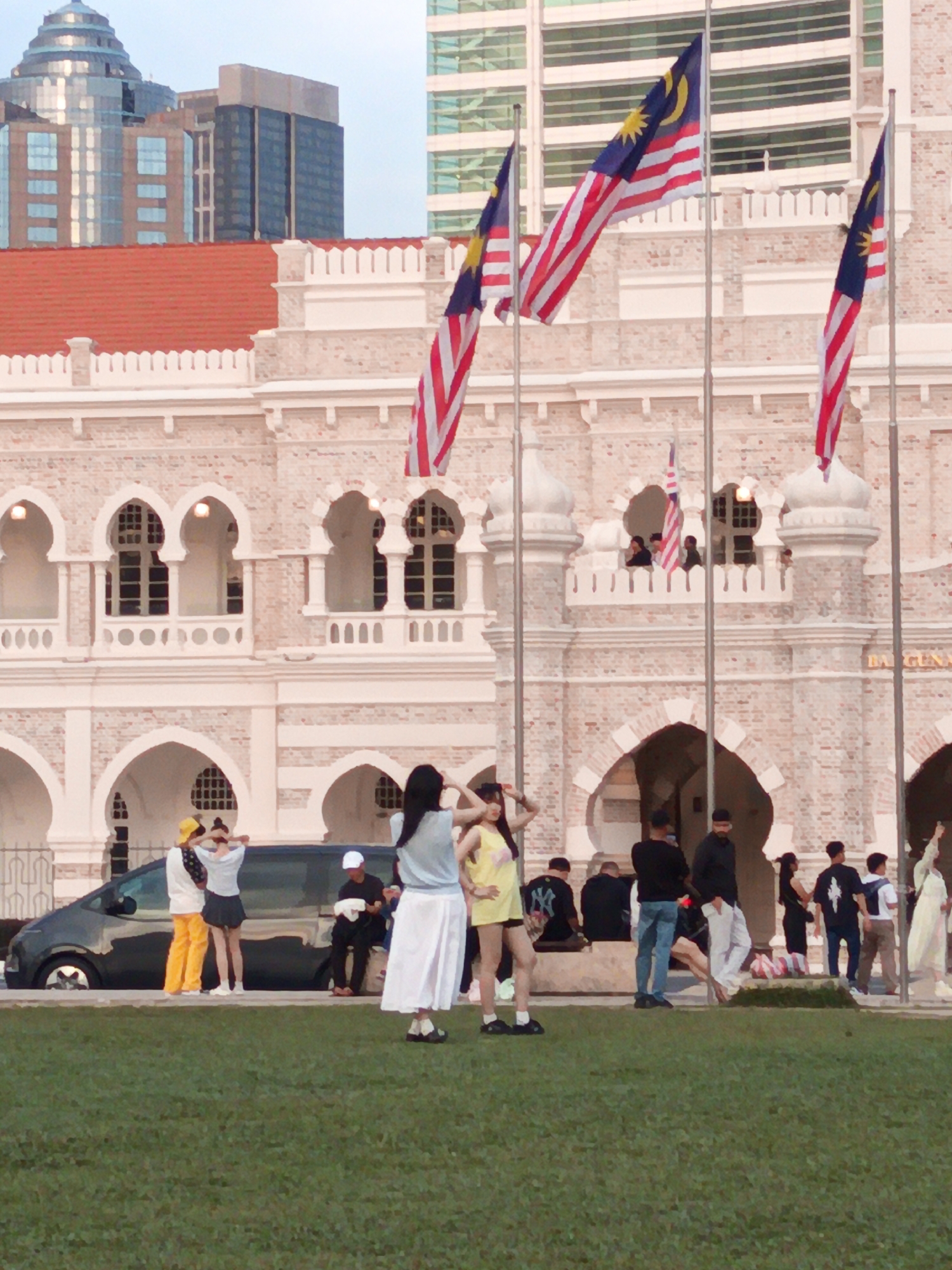 The image shows the Sultan Abdul Samad Building in Kuala Lumpur, Malaysia. The building is made of red brick with white arches and decorative elements. Several Malaysian flags are flying from poles in front of the building. In the foreground, a grassy area is visible, and several people are standing and walking around. To the left, a dark colored van is parked. The sky is overcast.