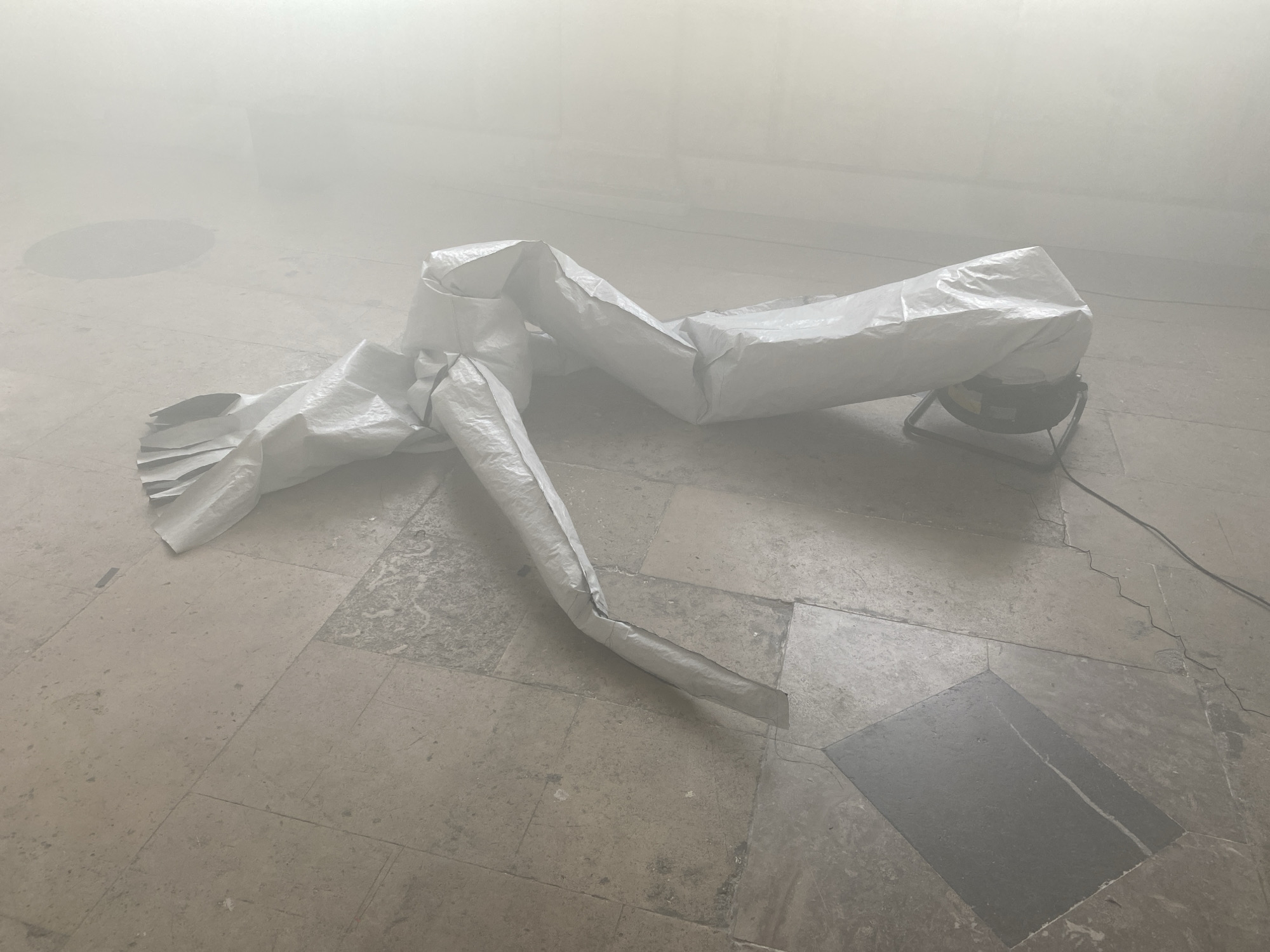 The image presents a foggy interior space with a tiled floor. A large, crumpled white tube, possibly made of plastic or fabric, stretches across the floor. One end of the tube is connected to a black fan or blower, which is supported by a metal frame. An electrical cord extends from the fan. The scene is obscured by a dense fog or haze. Based on the architecture of the floor, the location could potentially be a historic building. Without further information, pinpointing the location in Paris is not possible.