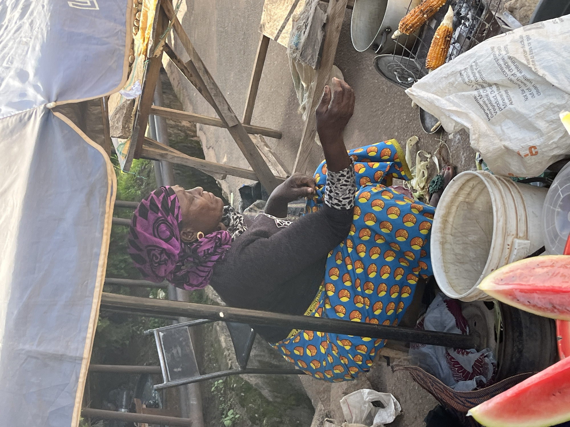 An elderly woman wearing a purple headscarf and a black top with a leopard print collar sits on a stool. She is dressed in a brightly colored blue and orange patterned dress. Her right arm is raised, with her hand open. Her left arm rests on her lap. She is in an outdoor setting, possibly a market. Behind her, a wooden structure supports various items, including a large white bag with visible text, a metal grate with corn on it, and a white bucket. Slices of watermelon are visible in the foreground. The scene appears to be during daylight. The visible text on the bag includes ingredients and warnings, such as "Not for human consumption." The overall impression is of a market stall in Barakin Akawo, Nigeria, where the woman might be selling goods.