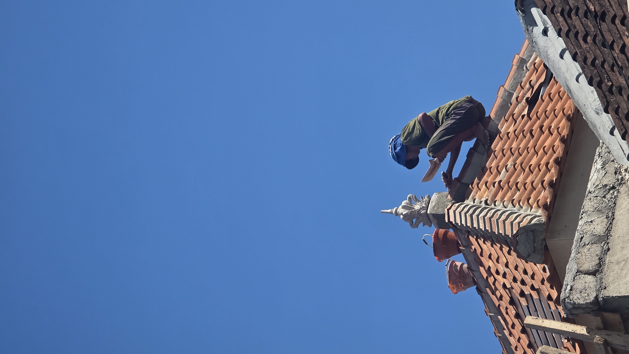 An individual wearing a blue cap and a green shirt is crouched on an orange tiled roof, working on its peak under a clear blue sky. The person appears to be a construction worker, possibly engaged in repair or maintenance. They are holding a tool, possibly a trowel or spatula, and their hands are positioned as if smoothing or applying a material. To the left of the worker, there are two orange buckets. One bucket is placed directly on the roof tiles, while the other is slightly behind it. A decorative architectural element, resembling a sculpted finial, is positioned near the peak of the roof, just behind the buckets. The roof itself is made of terracotta tiles, with some visible wooden support beams on the left side. The surrounding environment suggests an outdoor setting, likely in a residential or urban area, given the presence of rooftops. The image captures a moment of focused work on a bright, sunny day.