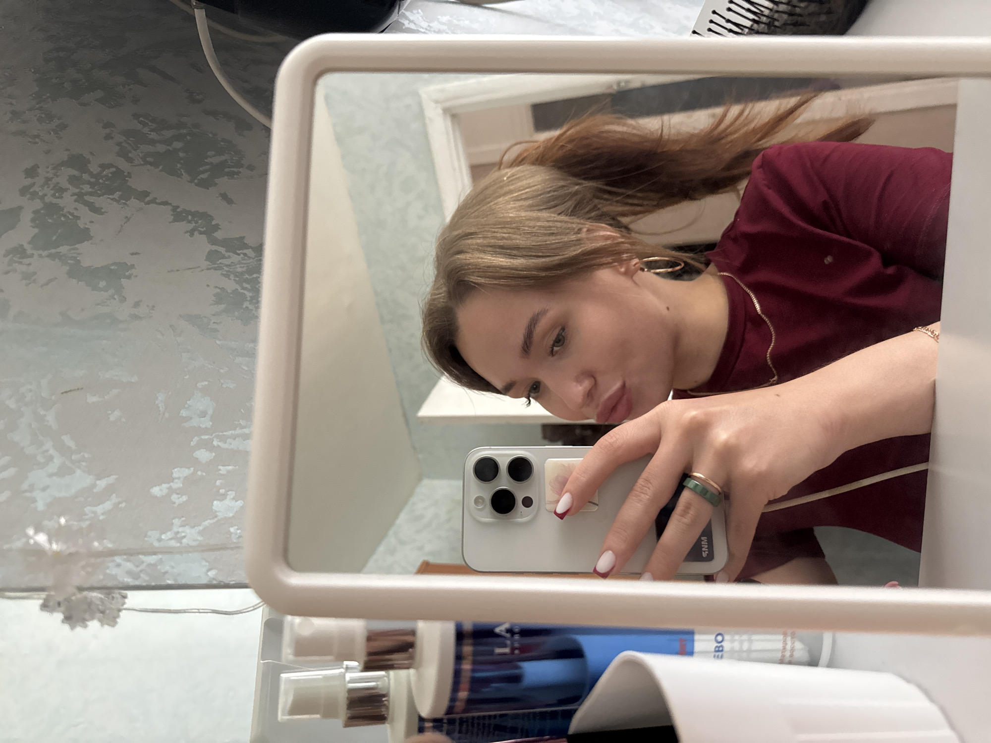 The media file shows a close-up reflection in a mirror of a young woman taking a selfie with her smartphone. She is holding the phone with her right hand, which has manicured nails with a French tip design and two rings on her finger, one of which appears to be jade. She is looking at her reflection with a slight pout.
The woman has long, light brown hair tied back in a ponytail and is wearing a dark red t-shirt with a thin gold chain. She has a large hoop earring in her left ear. The background of the reflection shows a room with a textured wall and a glimpse of what appears to be a light fixture or decoration.
In the foreground of the image, partially obscured by the mirror, are various objects that appear to be toiletries or cosmetic products. One bottle is visible with blue and white labeling, with the text "EBO" partially readable.
The overall scene appears to be a casual, indoor setting, likely a bathroom or dressing area. There are no explicit indicators of the time of day or weather conditions. The woman's expression suggests a playful or relaxed mood. The location context provided is Chaikovsky, Russia.