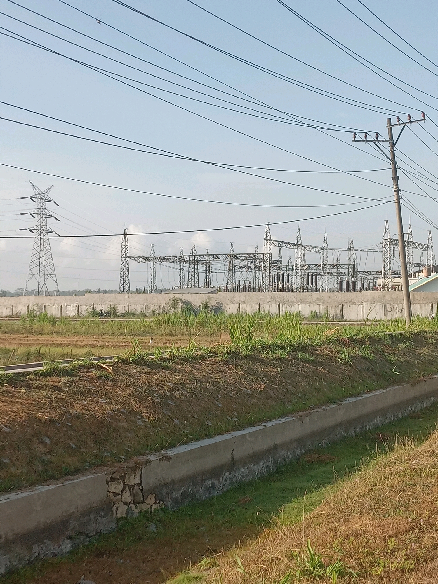 A medium shot captures an outdoor scene under a clear, light blue sky, likely during daylight hours. In the foreground, a narrow concrete drainage ditch runs horizontally across the lower portion of the frame, with dry, brown grass and some green vegetation on its banks. Beyond the ditch, a field of dry, brown soil and some patches of green grass stretches towards a low, concrete wall. Two figures are visible near the wall, one appearing to be riding a motorcycle. Behind the wall, a complex electrical substation dominates the middle ground. It features numerous metal towers, insulators, and wires, indicative of power transmission and distribution infrastructure. To the left of the substation, a tall, lattice metal pylon stands prominently. The background consists of more of the clear sky, with a few wispy clouds. The overall environment appears rural or semi-rural, with evidence of agricultural land and extensive electrical infrastructure, suggesting an area near industrial or power-generating facilities. The scene is located in the area of Temon, Indonesia.