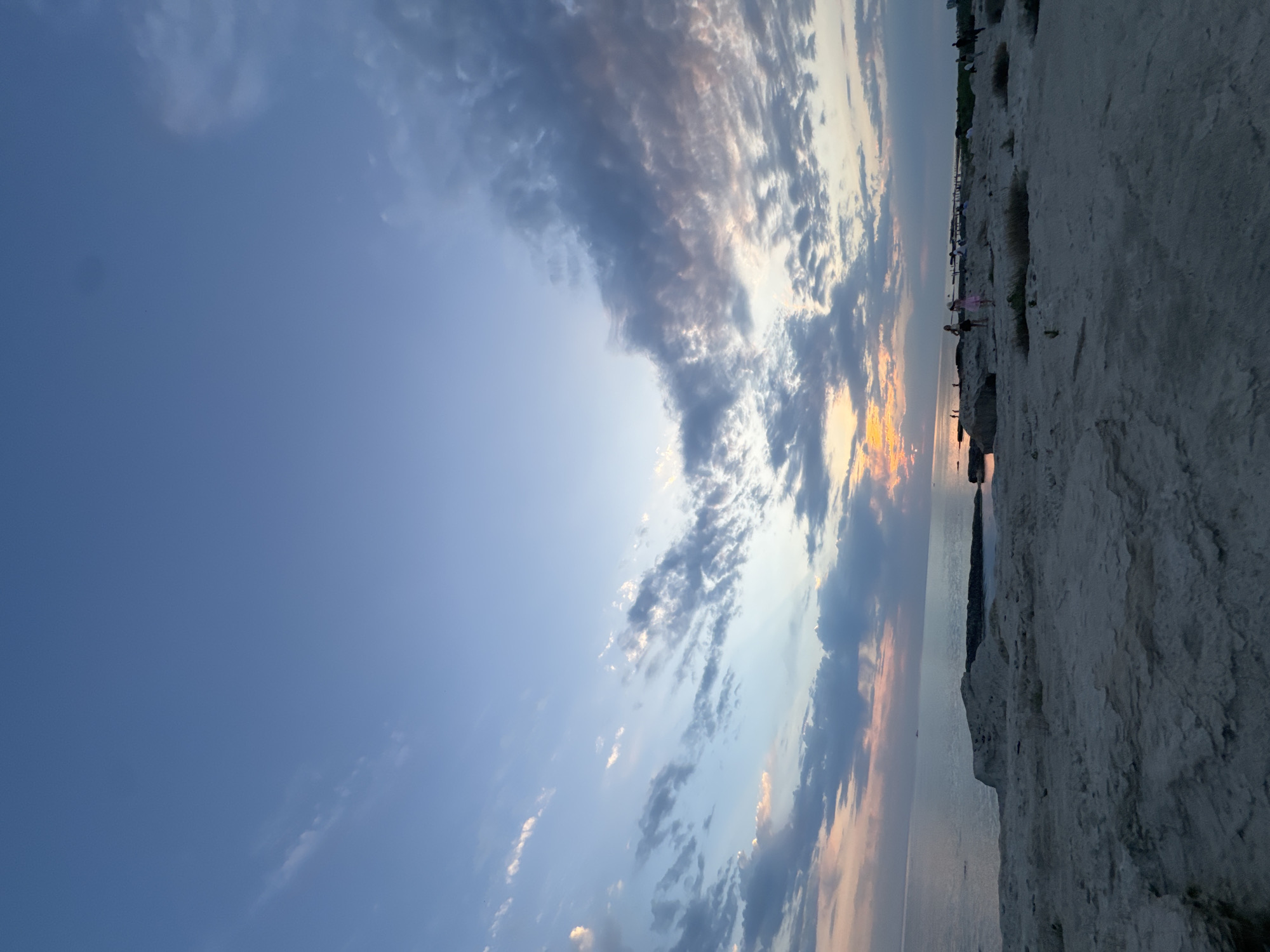 An outdoor, eye-level, wide-angle shot captures a coastal landscape at sunset, with the Caspian Sea stretching towards the horizon under a vast sky. The foreground is dominated by a textured, light-colored rocky terrain. In the middle ground, the sea meets the land, with several large rock formations scattered along the coastline. A few small figures, appearing to be people, are visible on the rocky expanse and near the water's edge. The sky is filled with scattered clouds, tinged with the warm hues of the setting sun, transitioning to blues and grays as it extends upwards. The overall scene suggests a serene, natural environment. This location is the coast of Aktau, Kazakhstan.