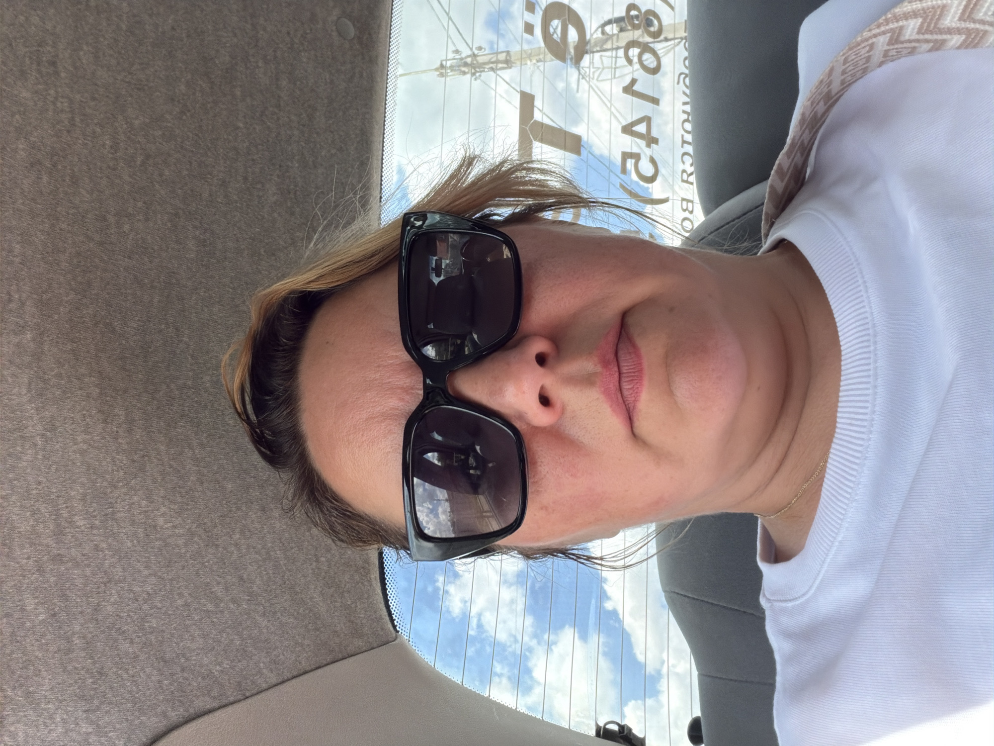 The image shows a medium shot of a woman wearing large, black-rimmed sunglasses and a white crewneck sweatshirt, seated in the back of a vehicle. Her hair is pulled back and blonde strands frame her face. Her expression is neutral, with her lips slightly parted. The interior of the car is visible, including a gray headrest and the fabric of the car's ceiling. Through the rear window, a bright blue sky with scattered white clouds is visible, along with a utility pole and some text printed on the glass. The text appears to be a company name, "Tē" and "BO ROTONYADS" which are likely translated from Russian. The woman's face is the primary subject of the image.