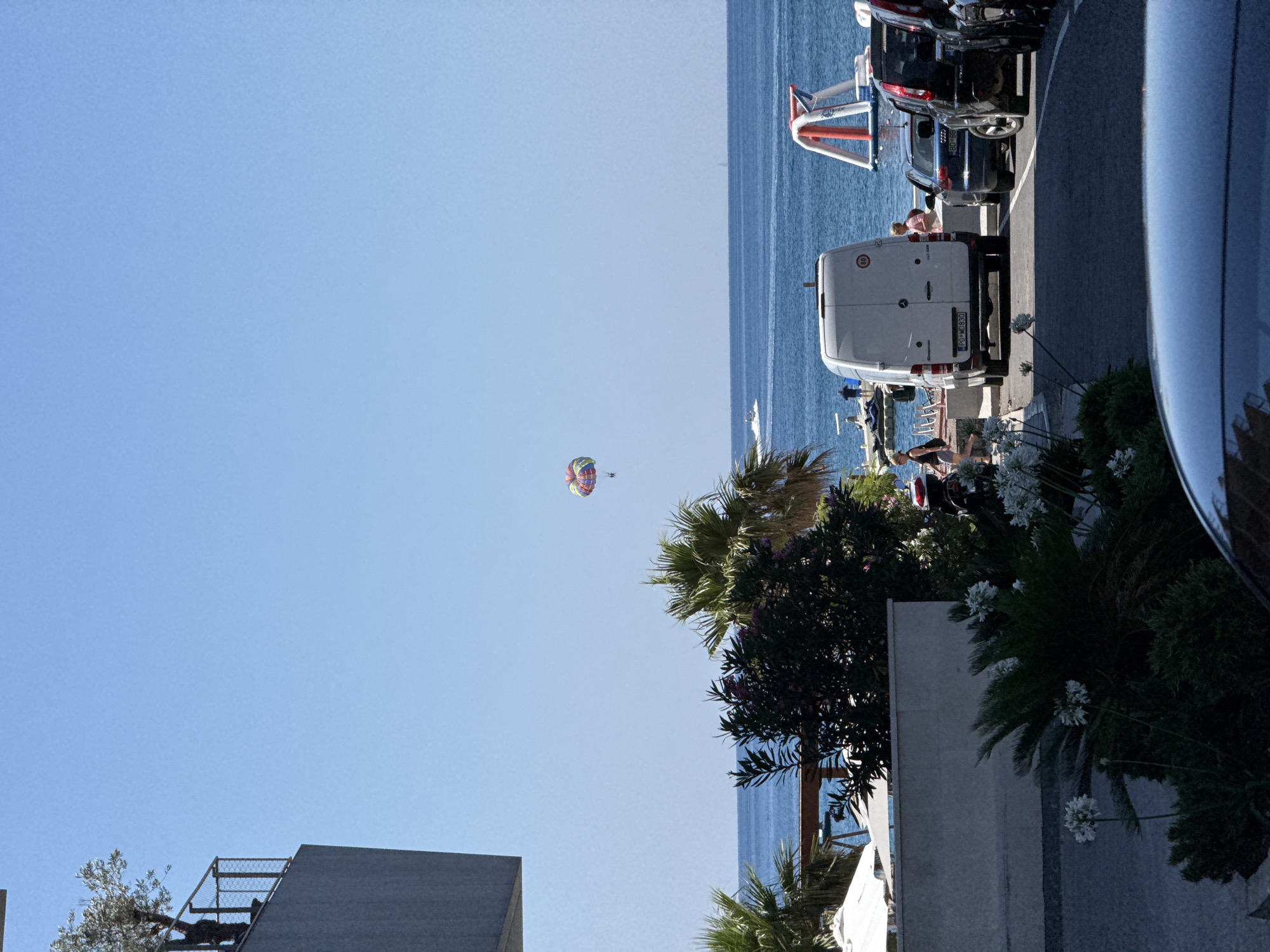 The image depicts a sunny day scene in Boreti, Montenegro, likely near a coastal resort. In the sky, a colorful parachute with a person suspended beneath it is visible, engaged in parasailing over the blue sea. The sea itself is calm, with a small boat or jet ski visible in the distance.
On the shore, a white van is parked, with a speed limit sign (80) on its rear. Several cars are parked behind the van, including a dark blue sedan and a black SUV. A woman in a pink top is standing near the van. Further back, there is an inflatable water attraction with red and white elements in the sea.
To the left of the scene, there is a modern building with a grey facade and a balcony featuring an olive tree. Lush green vegetation, including palm trees and flowering bushes, is prominent in the foreground and mid-ground, suggesting a well-maintained outdoor area. White umbrellas or canopies are also visible, indicating a beach or relaxation area. The ground in the foreground appears to be a paved walkway or parking area, with a dark car partially visible in the bottom frame.