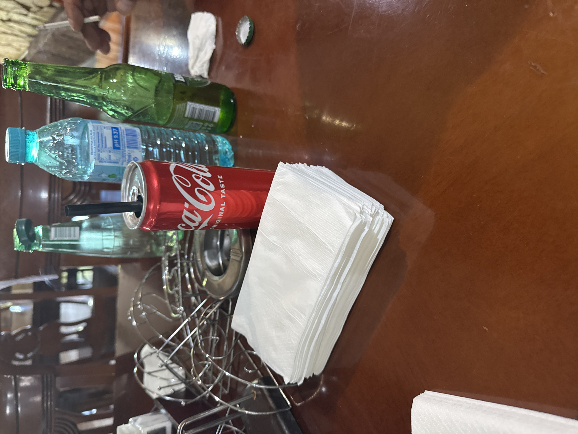 The image captures a close-up view of a wooden table, likely in a restaurant or dining area, with several items prominently displayed. In the foreground, a stack of white paper napkins is neatly arranged. To the right of the napkins sits a red Coca-Cola can with a black straw inserted. Behind the can, a clear plastic bottle of water with a blue cap is visible, and further to the right, a green glass bottle, likely containing a beverage, is placed. A white paper napkin is crumpled on the table near the green bottle, and a green bottle cap is also present. In the background, out-of-focus furniture, possibly chairs, can be observed. There is no visible indication that this scene is located in the city of Sredets, Bulgaria.