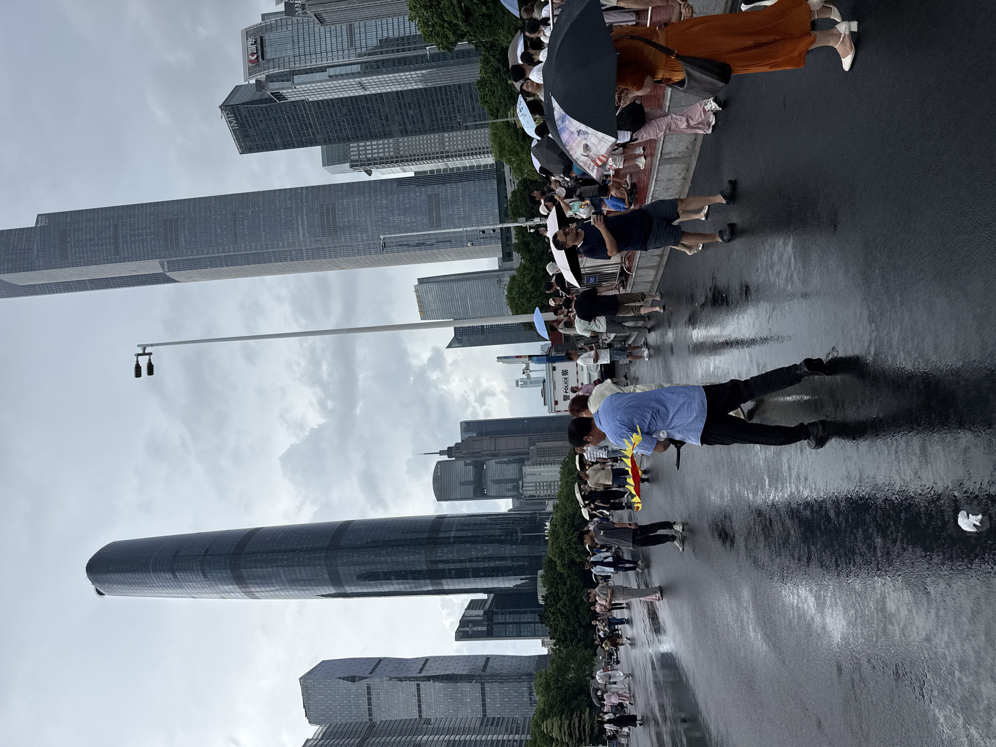 In Tianhe District, China, under a cloudy sky, a crowd of people stand on a wet, reflective paved area, suggesting recent rain. Many individuals are holding umbrellas, indicating a damp environment. In the background, several modern skyscrapers dominate the skyline, characterized by their sleek glass facades and imposing height. In the foreground, a man in a blue collared shirt and black pants stands with his back to the camera, holding a yellow and red object. To his right, a woman in an orange dress walks away, carrying a dark handbag. Other people are scattered throughout the scene, some walking, some standing, and some interacting with each other, while a white police vehicle is visible on the right side of the frame.