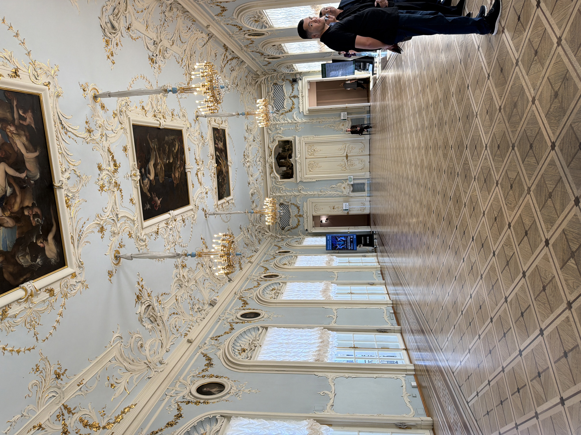 The image depicts the interior of a grand ballroom in the Catherine Palace, located in Pushkin, a municipal town within the federal city of Saint Petersburg, Russia. The ballroom features an ornate, pale blue ceiling adorned with intricate white stucco molding and gilded foliage. Two large oil paintings are set into the ceiling, one above the other, and several ornate chandeliers hang from it. The walls are also pale blue and decorated with similar stucco work, with tall, arched windows along one side covered by sheer white curtains. The floor is a highly polished wooden parquet in a diamond pattern.
In the foreground, on the right side of the frame, stand three men. The man closest to the camera is wearing a black polo shirt and dark jeans, with black sneakers. To his right, two other men are partially visible, one in a black jacket and dark pants. They appear to be observing the room.
In the middle ground, several people are present. A woman sits on a chair near a large double door. A digital display board is positioned near the center of the room. Further down the hall, through an open doorway, other individuals can be seen. The overall impression is of a formal, historic, and well-maintained interior.