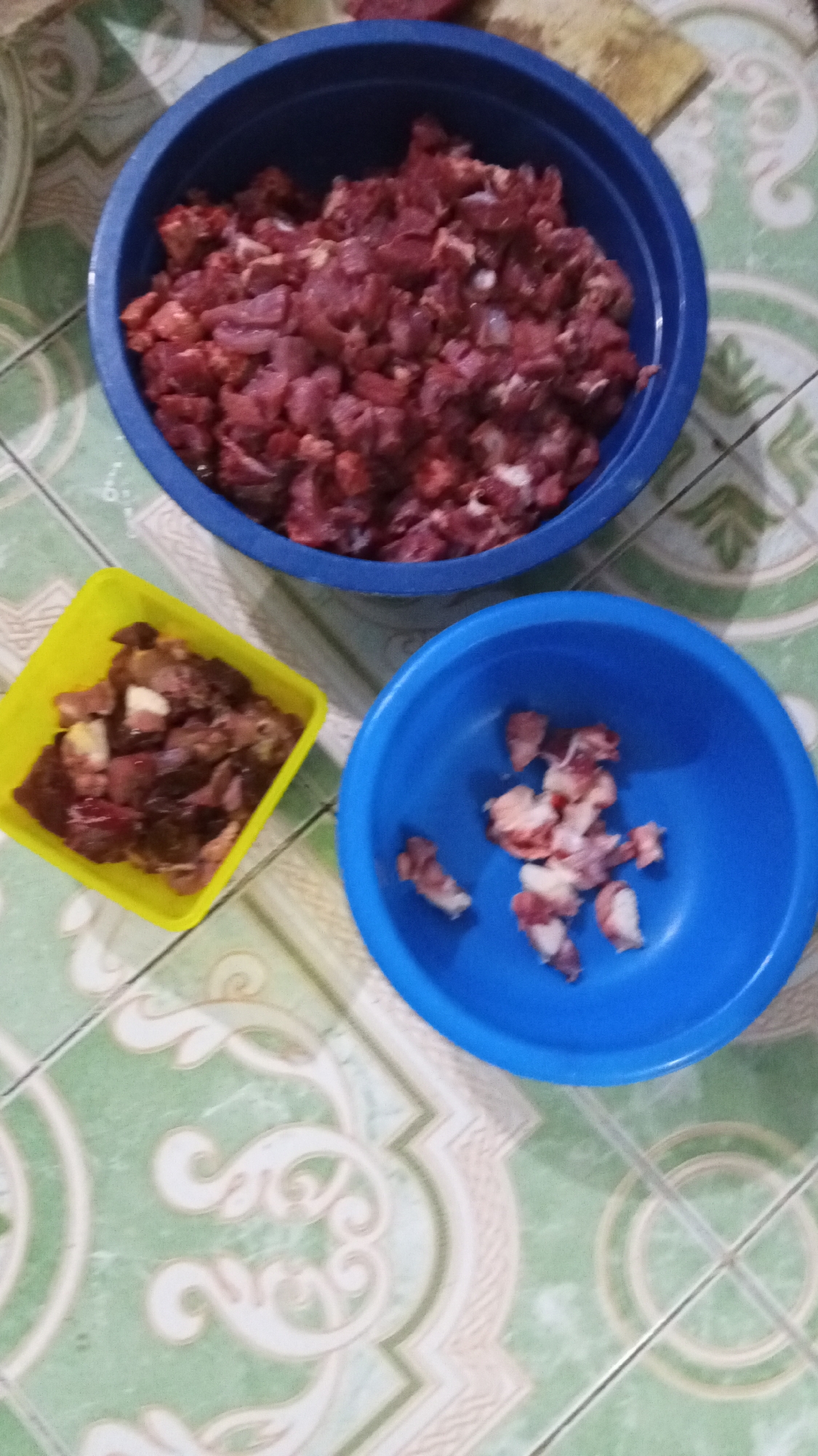 On a light green tiled surface with repeating stylized floral patterns, there are three containers holding raw meat. The largest container is a dark blue bowl filled with small, roughly diced pieces of red meat. To the left, a yellow square container holds larger, irregularly shaped pieces of meat, some darker in color. A smaller, light blue bowl to the right contains only a few pieces of meat, appearing to be mostly fat and connective tissue. The top corner of a cutting board is visible in the upper right. The scene likely depicts food preparation. The location is Lhokseumawe, Indonesia.
