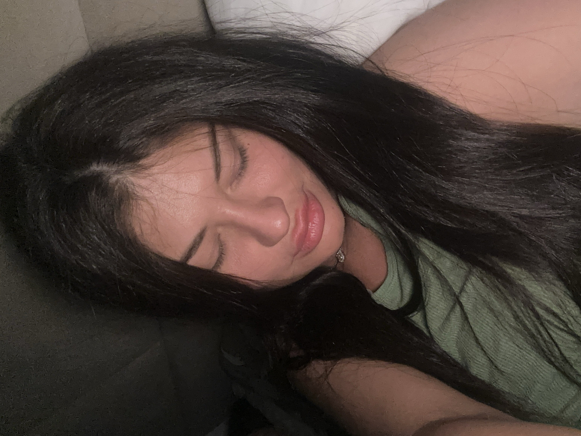 A young woman with long, dark hair lies with her eyes closed. She appears to be resting or sleeping. The woman is wearing a light green t-shirt. A white pillow can be seen in the background. The setting seems to be indoors, possibly in a bedroom. There is no information that can confirm if the location is in the city of Moscow, Russia.